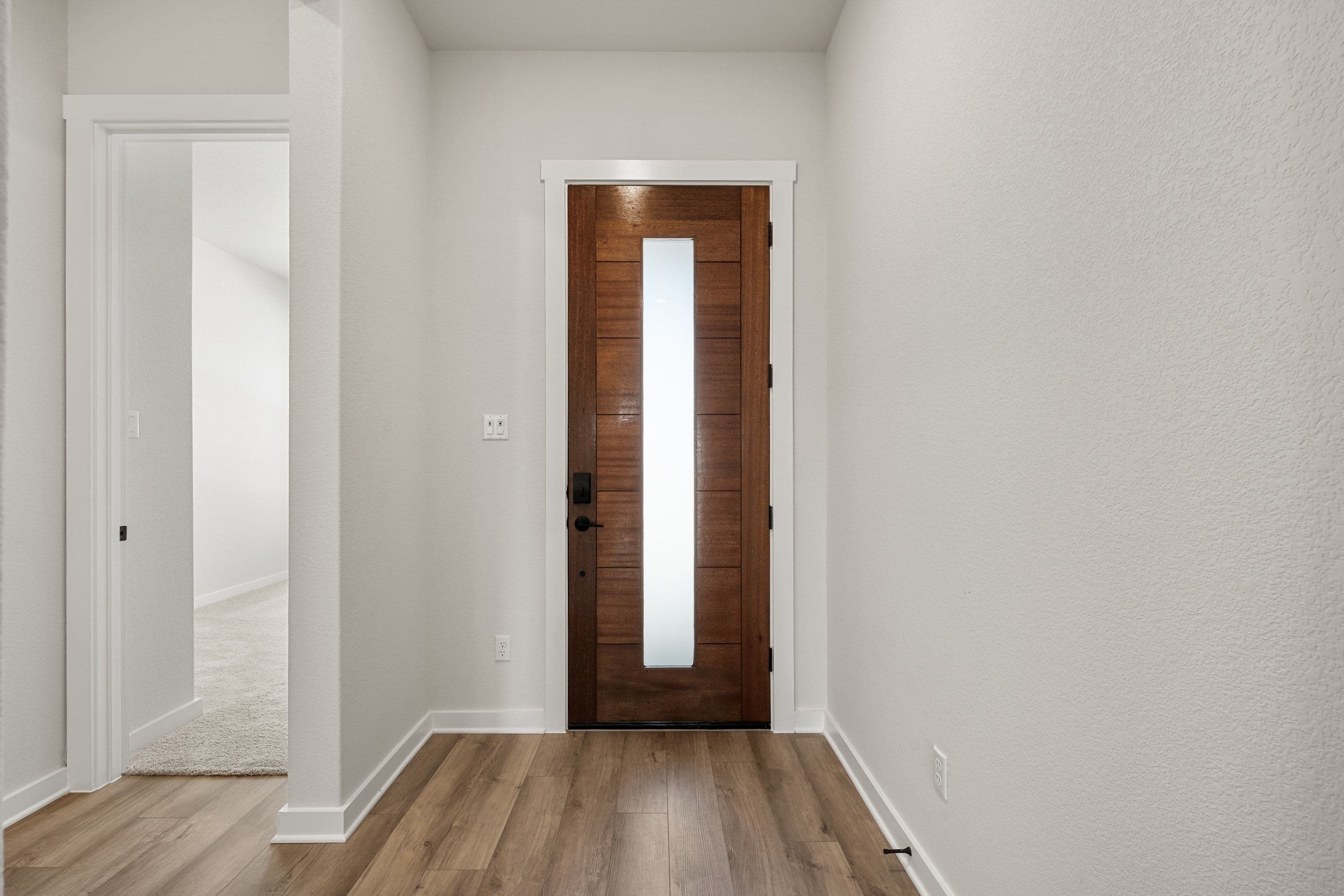 A modern wooden front door with a glass panel is centered in the foreground, with a hallway visible through an open doorway to the left, all set against plain white walls.