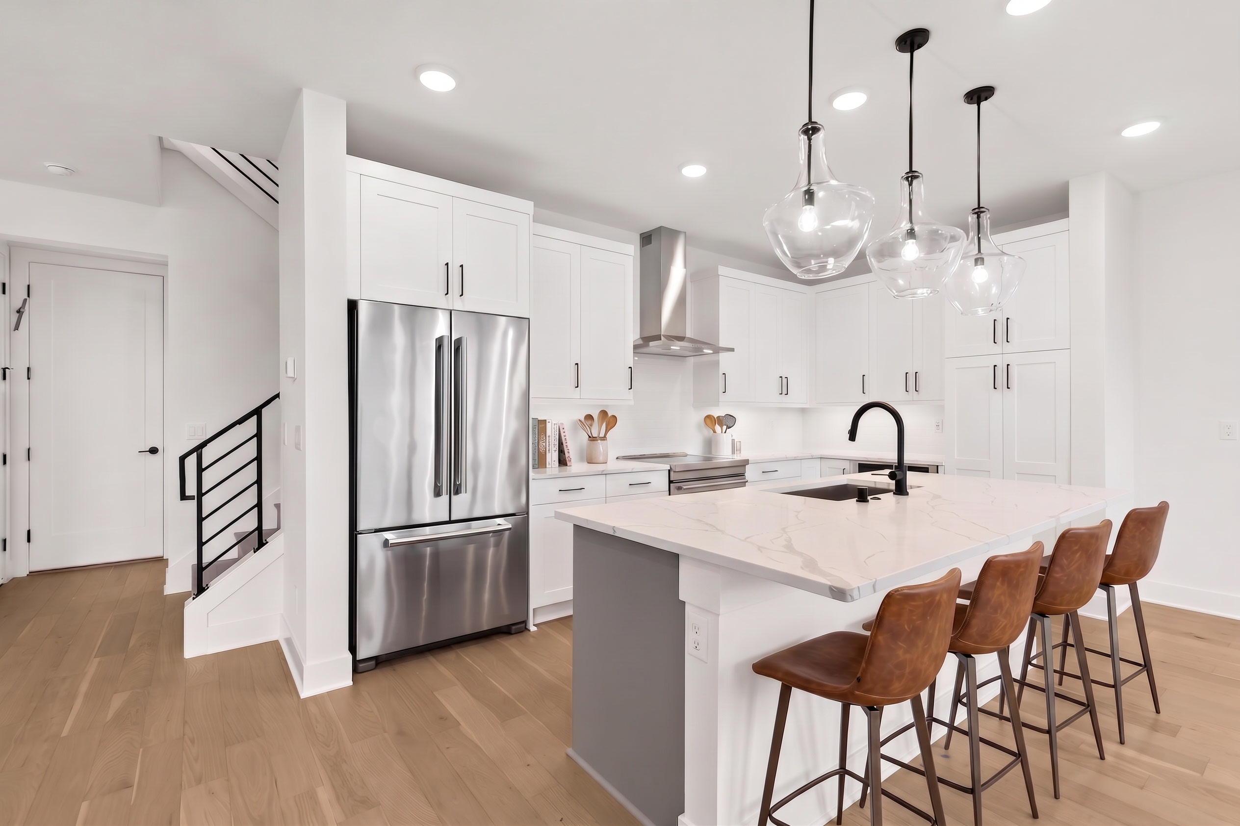 A modern kitchen with a stainless steel refrigerator and island with bar stools in the foreground, and white cabinets and a staircase in the background.