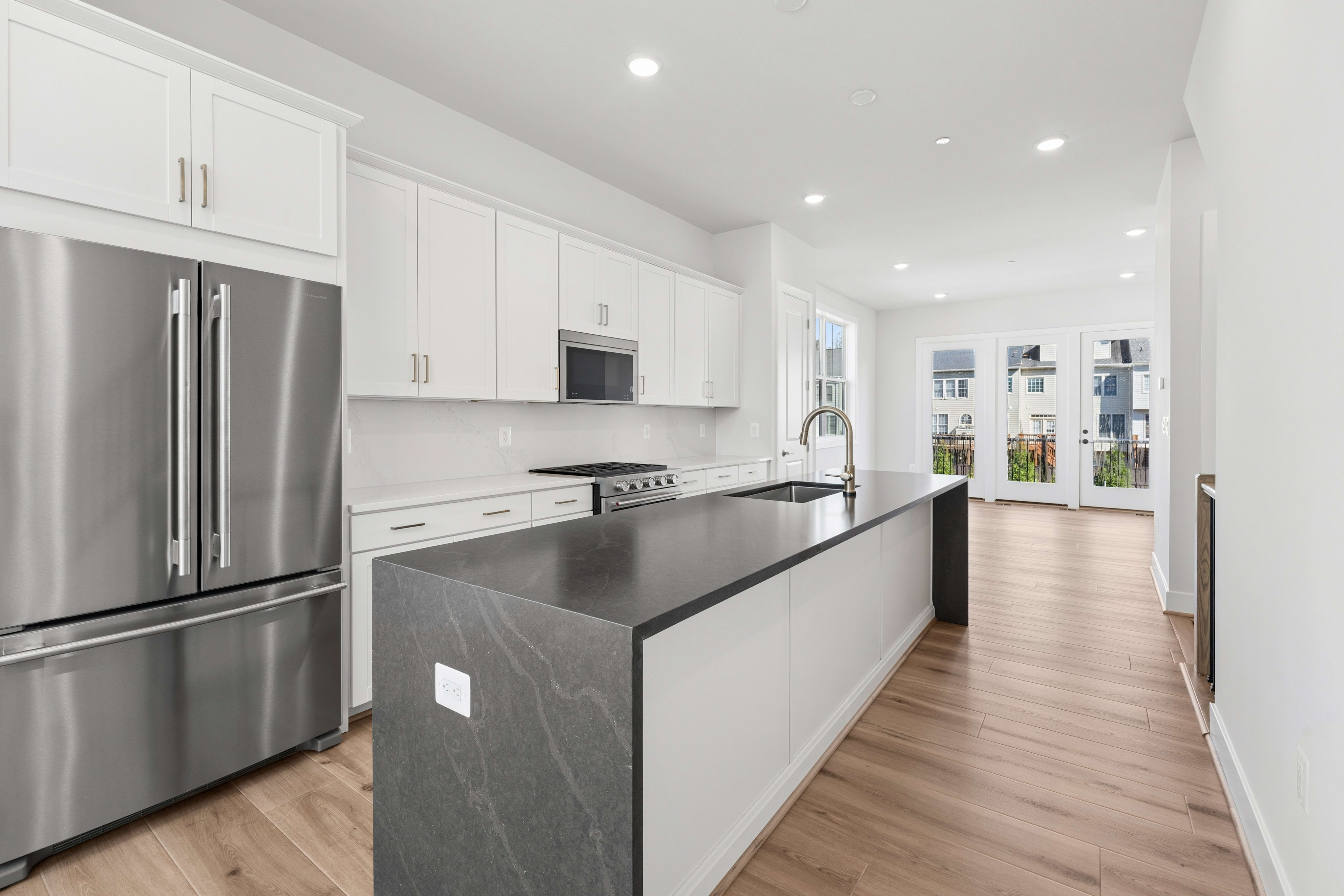 A modern kitchen with a large island in the foreground opens to a bright living area with double doors in the background.
