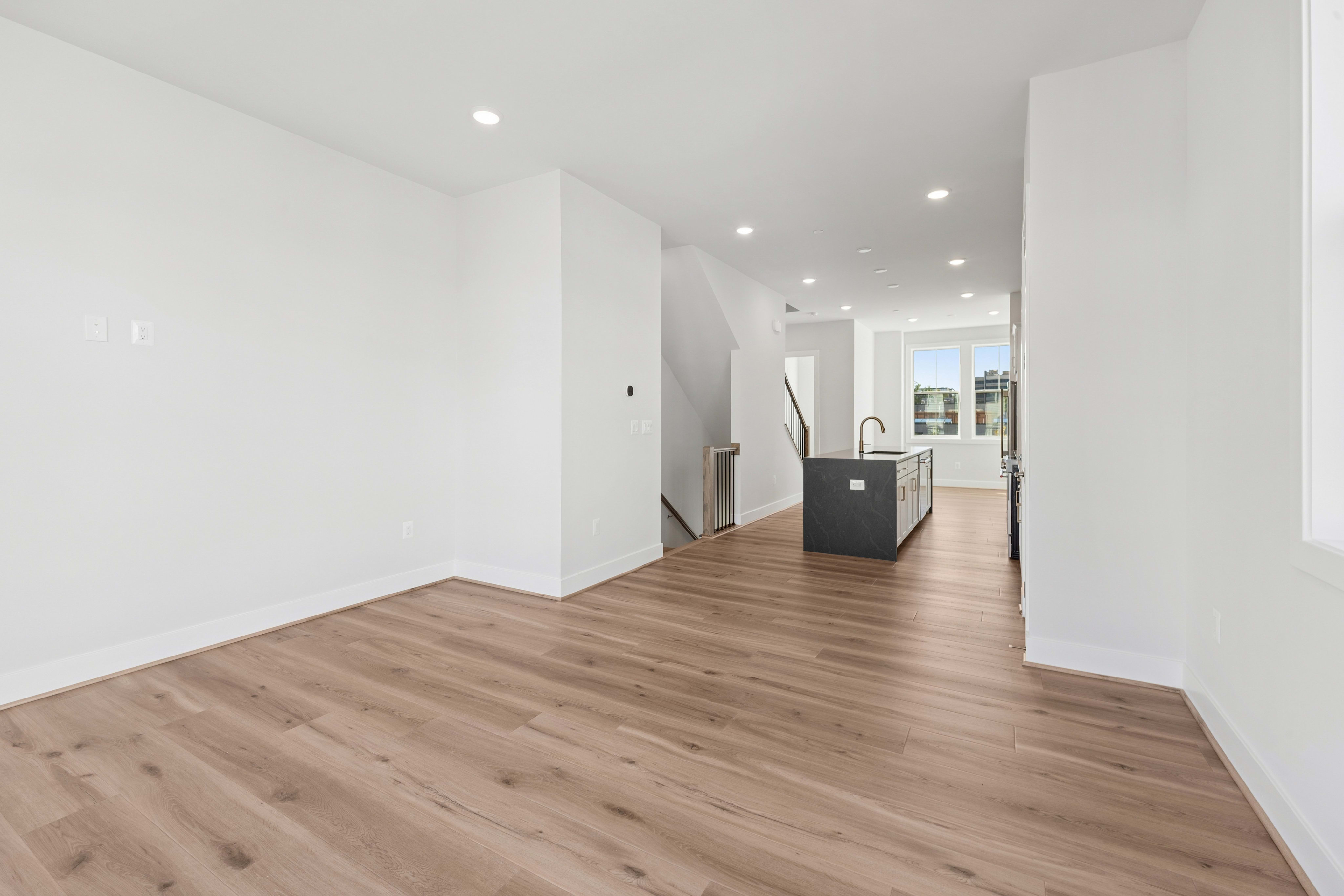 A modern living space with light wood flooring in the foreground opens to a kitchen area with a dark island and white cabinets in the background.