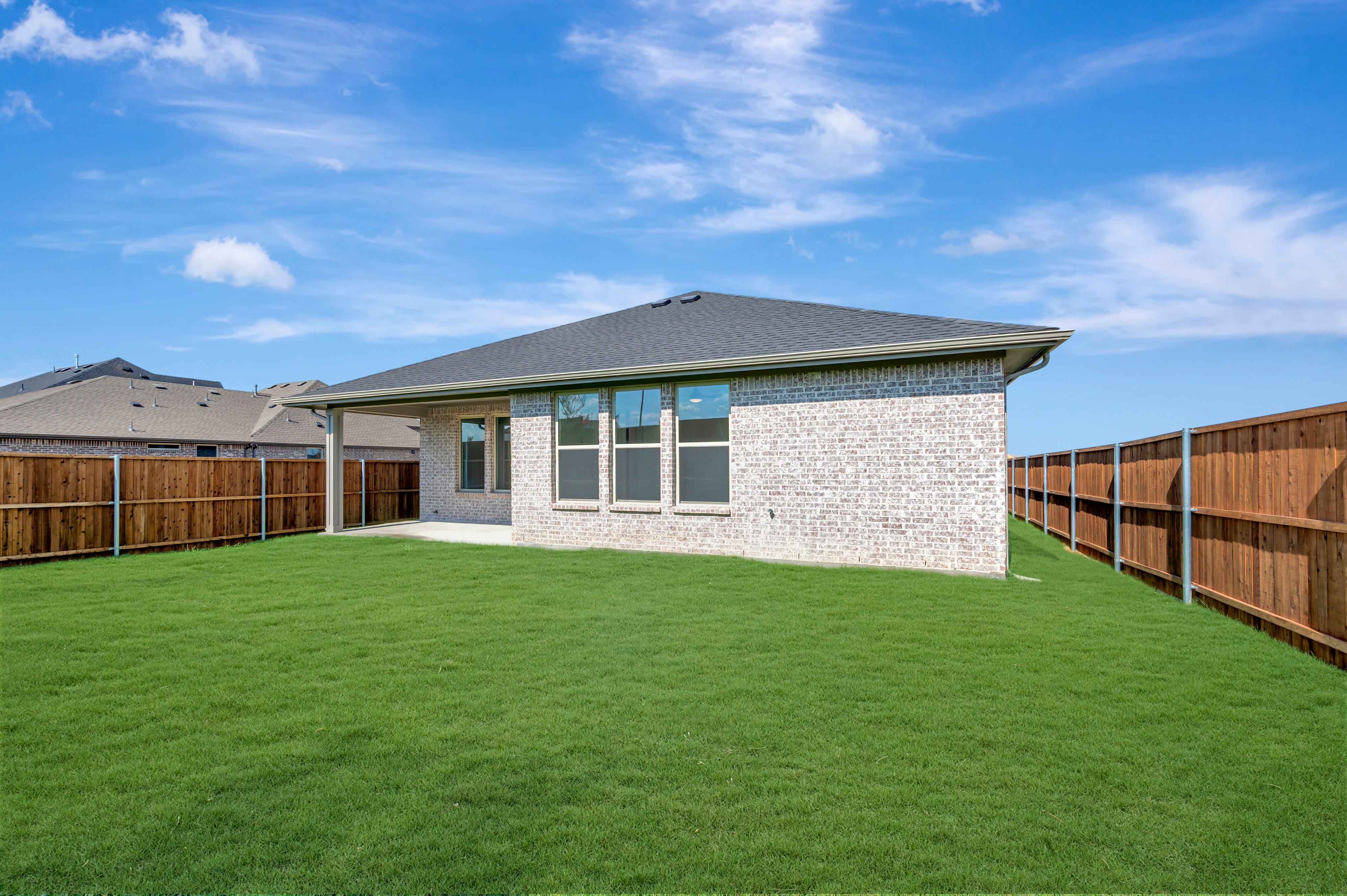 A freshly mowed green lawn fills the foreground, leading to a white brick house with a covered patio and a wooden fence enclosing the backyard under a bright blue sky with scattered clouds.