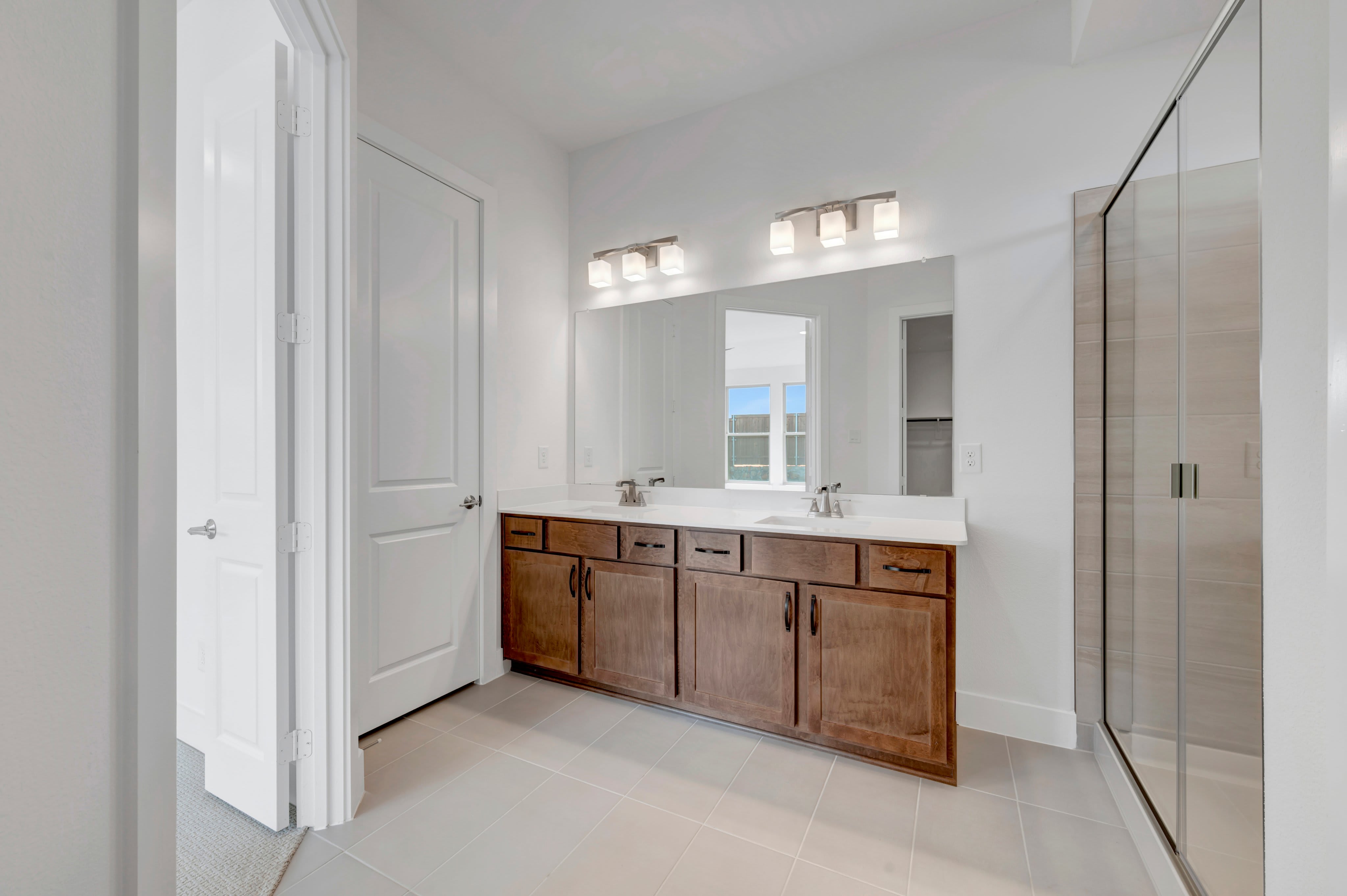 A modern bathroom features a double vanity with a large mirror in the foreground, and a glass-enclosed shower in the background.