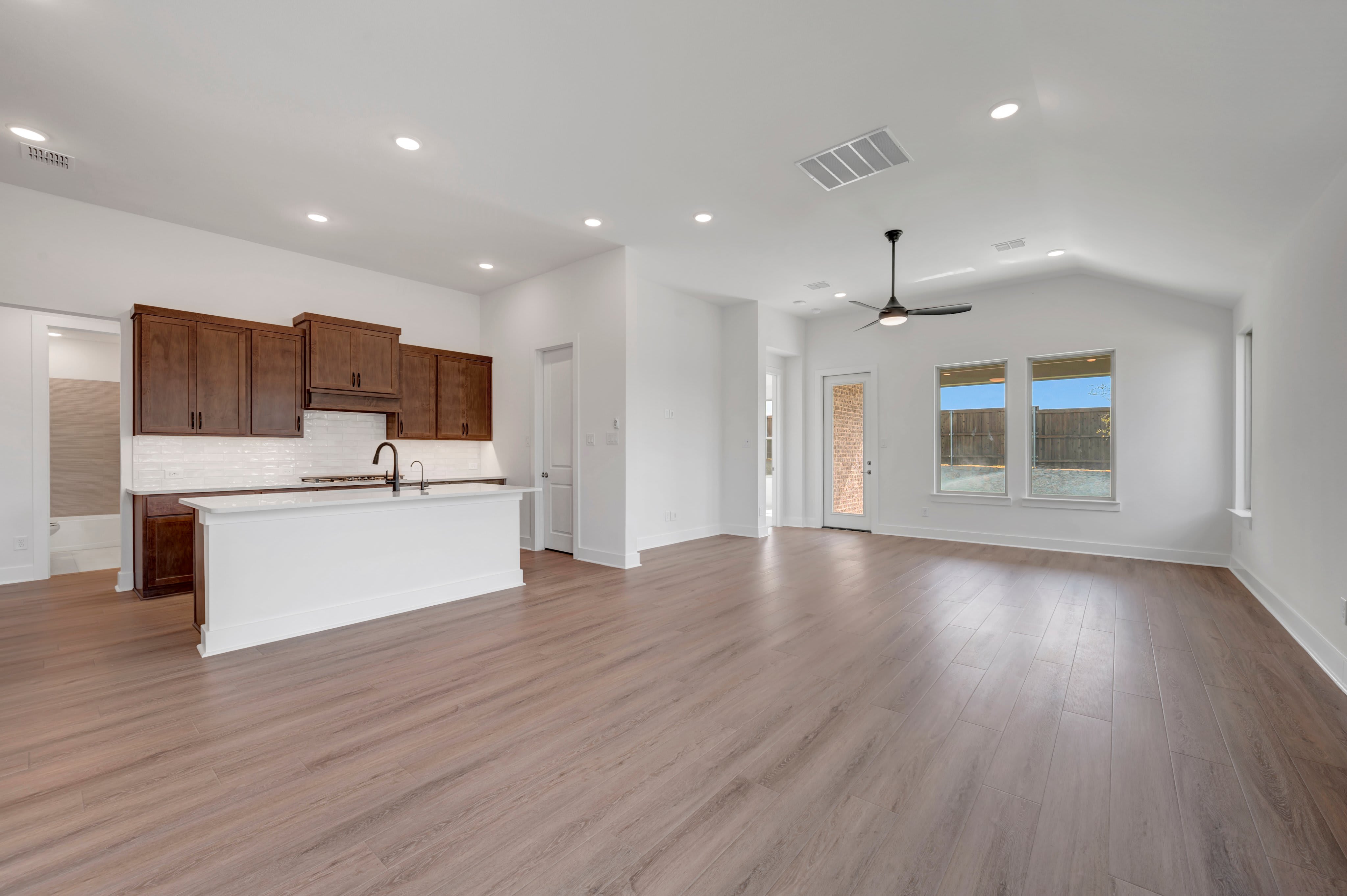A modern kitchen with dark wood cabinets and a white island is in the foreground, while a spacious living area with a vaulted ceiling and large windows overlooking a blue sky and trees is in the background.