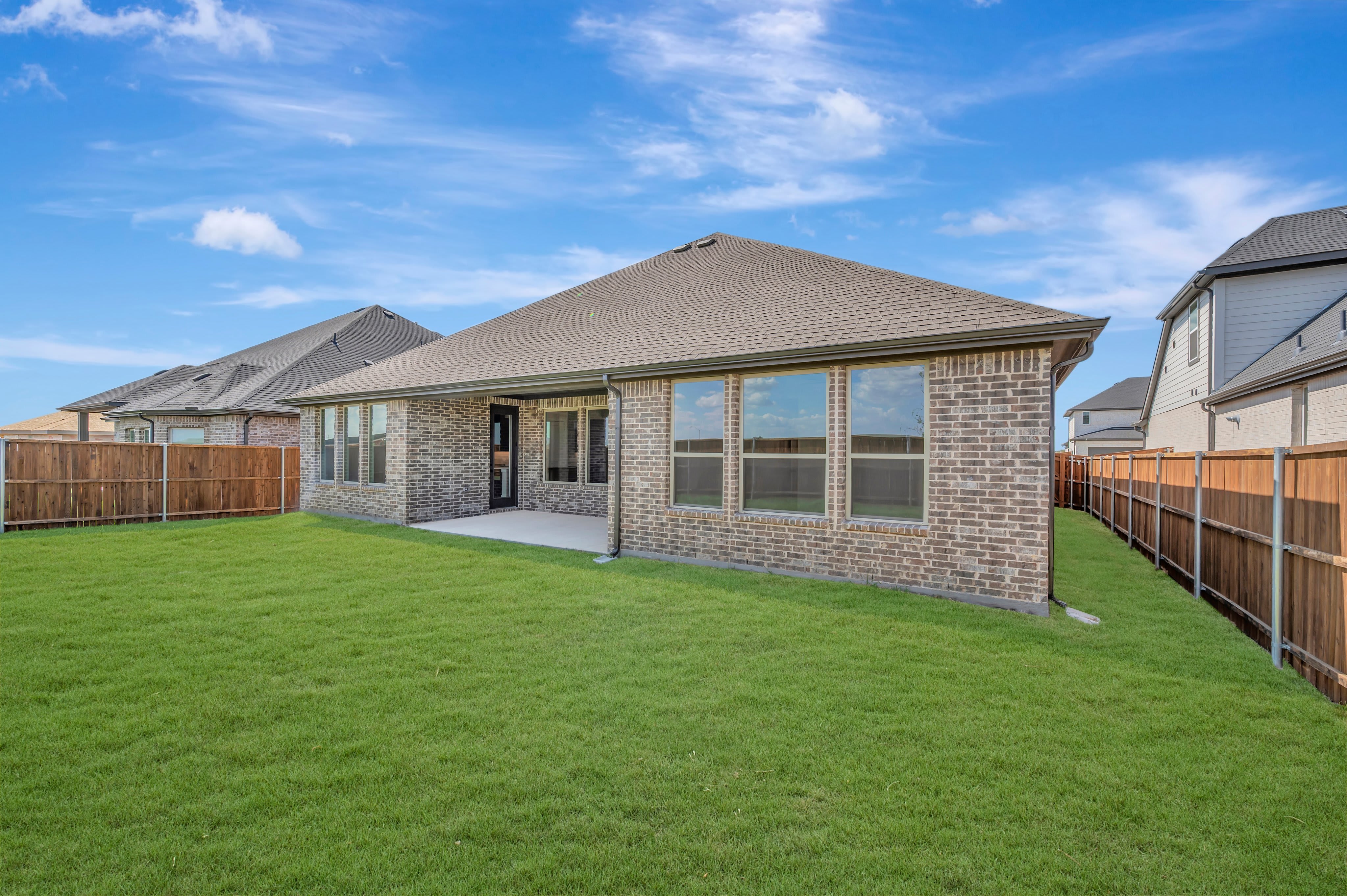 A freshly mowed green lawn stretches out in the foreground, leading to a brick house with large windows and a covered patio, all set against a backdrop of a bright blue sky with scattered clouds and neighboring houses.