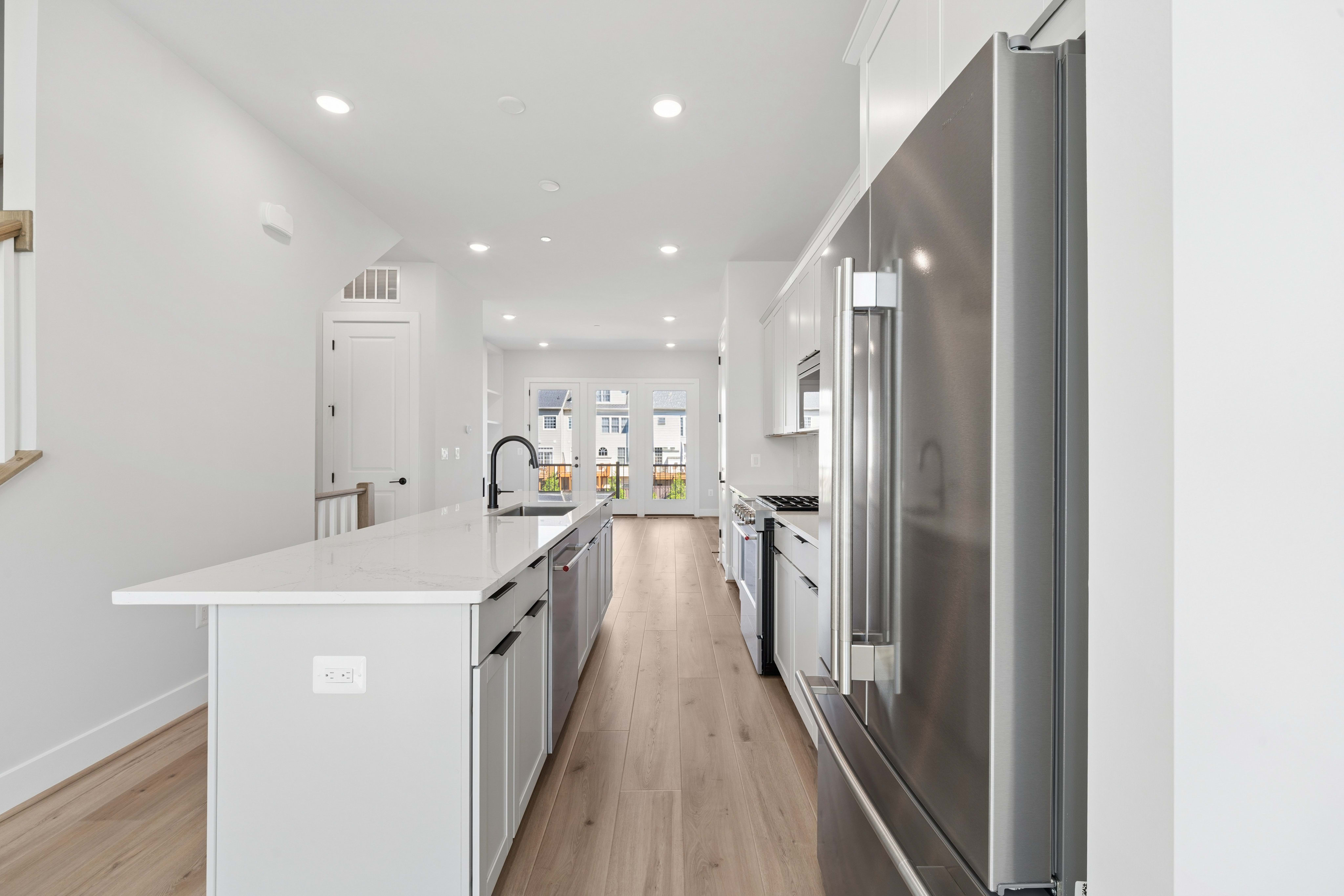 A modern kitchen with a white island and stainless steel appliances opens into a bright living area with large windows.