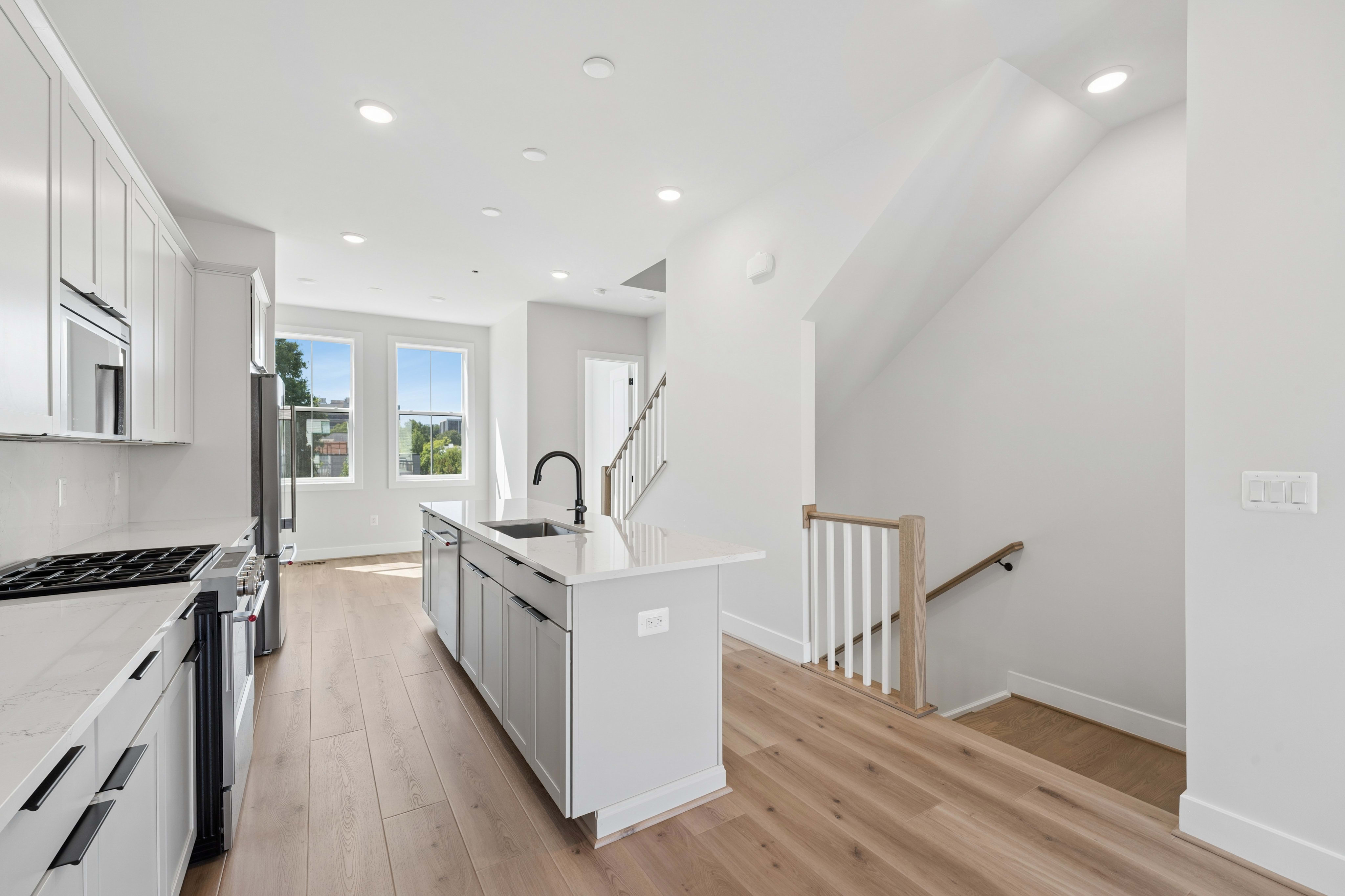 A modern kitchen with a white island and light wood floors is in the foreground, while a staircase and windows overlooking greenery are in the background.