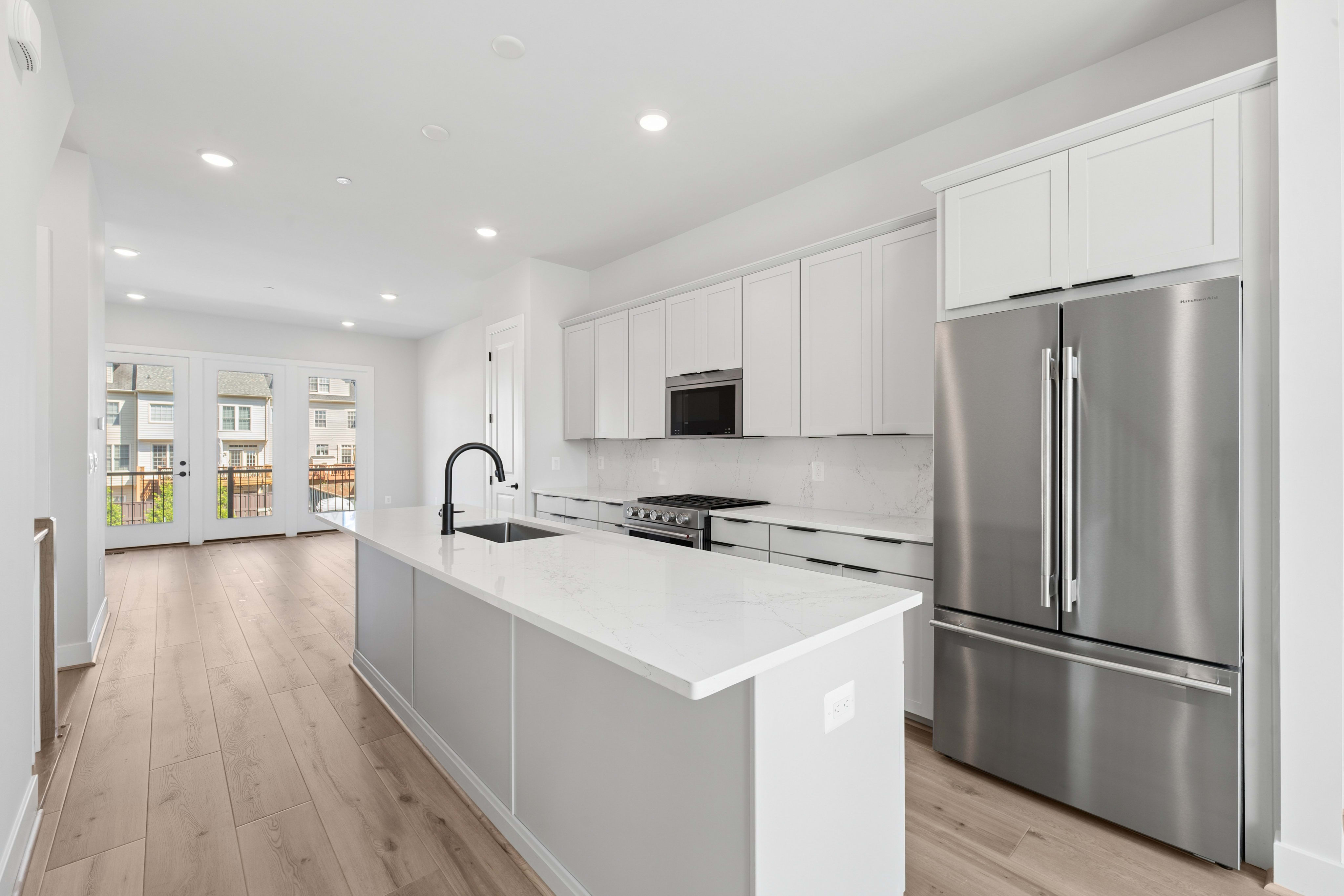 A modern white kitchen with a large island in the foreground opens to a living area with glass doors in the background.