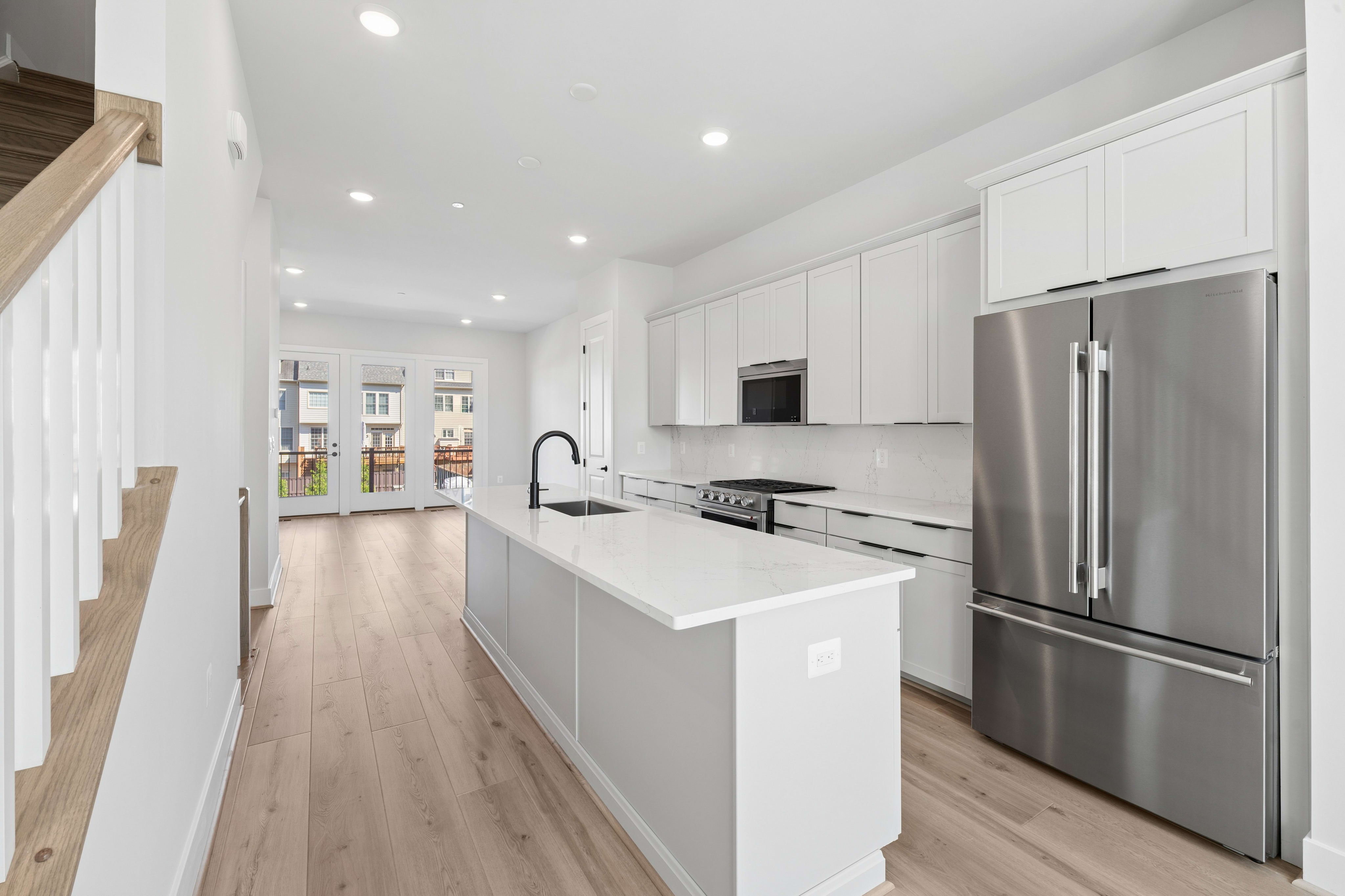 A modern white kitchen with a large island in the foreground and a living area with glass doors leading outside in the background.