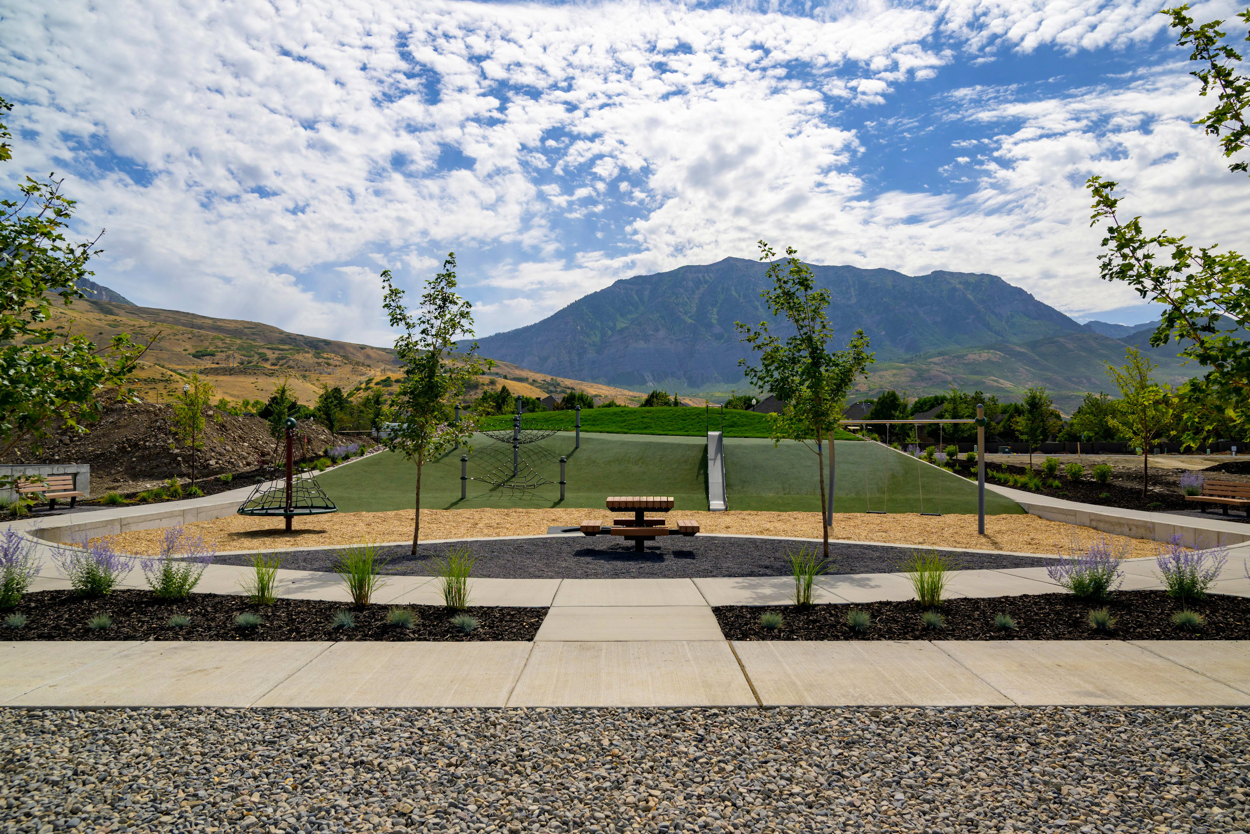A picnic table sits in the foreground of a park with a playground and a large mountain in the background under a cloudy sky.