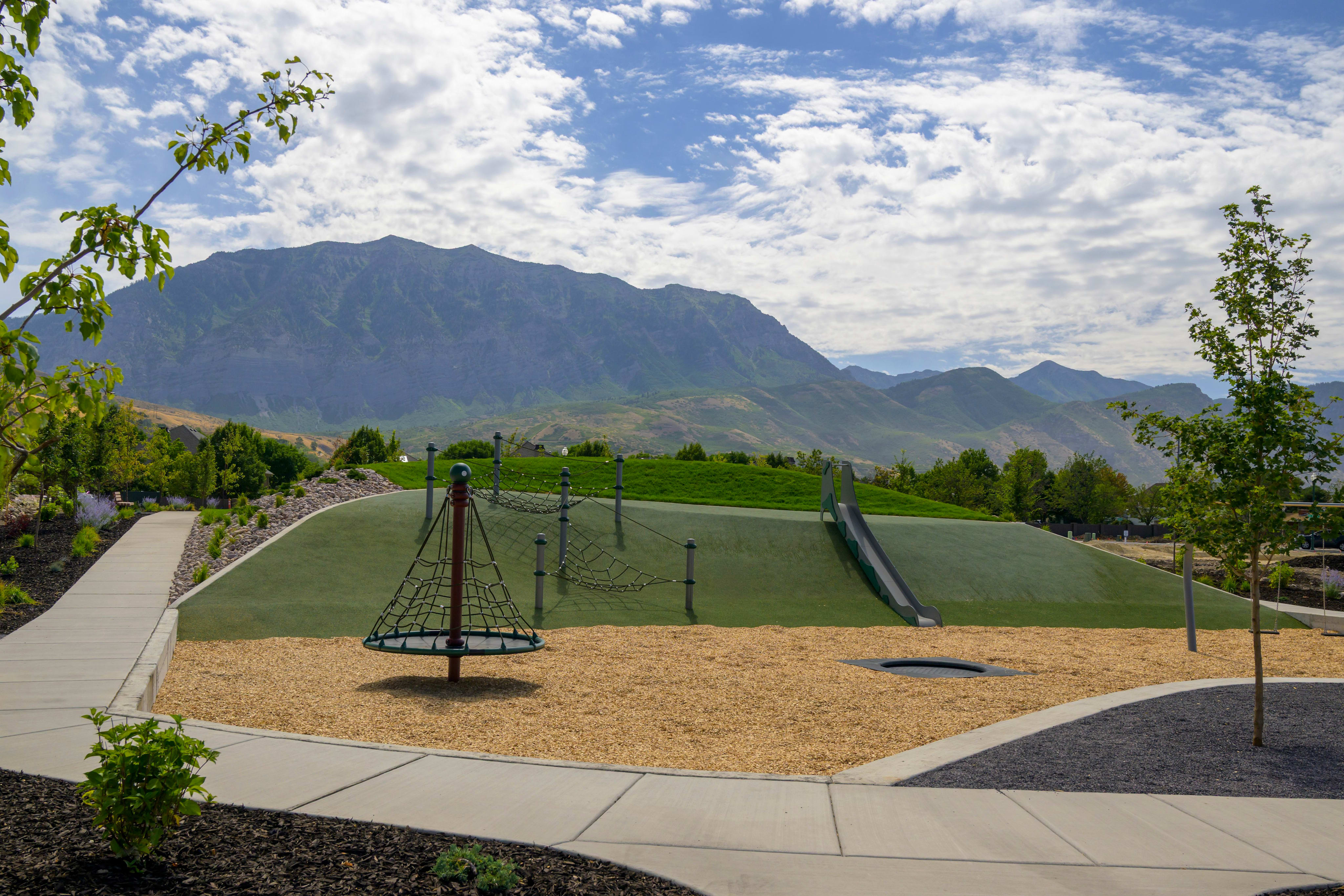 A modern playground with a spinning net in the foreground sits before a lush green hill and a majestic mountain range under a cloudy sky.