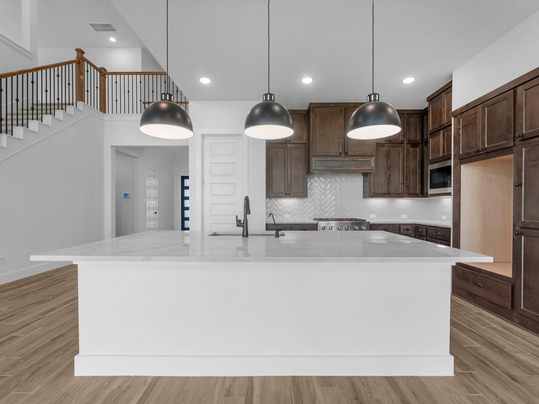 A modern kitchen island with pendant lights in the foreground contrasts with dark wood cabinetry and a staircase in the background.