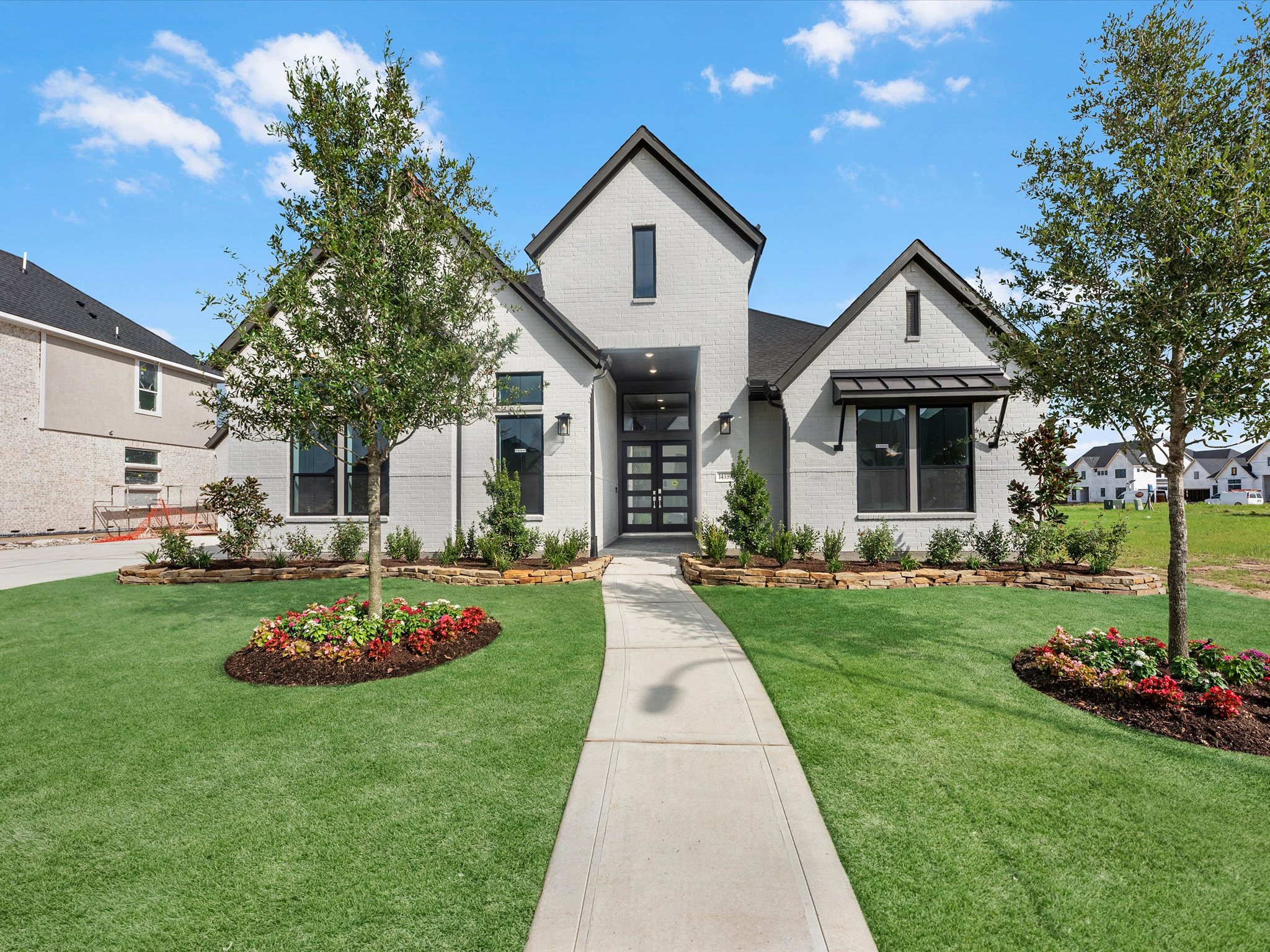 A modern white brick house with a gray roof and black trim sits on a manicured lawn with a concrete walkway leading to the front door, while other houses and a clear blue sky with scattered clouds form the background.