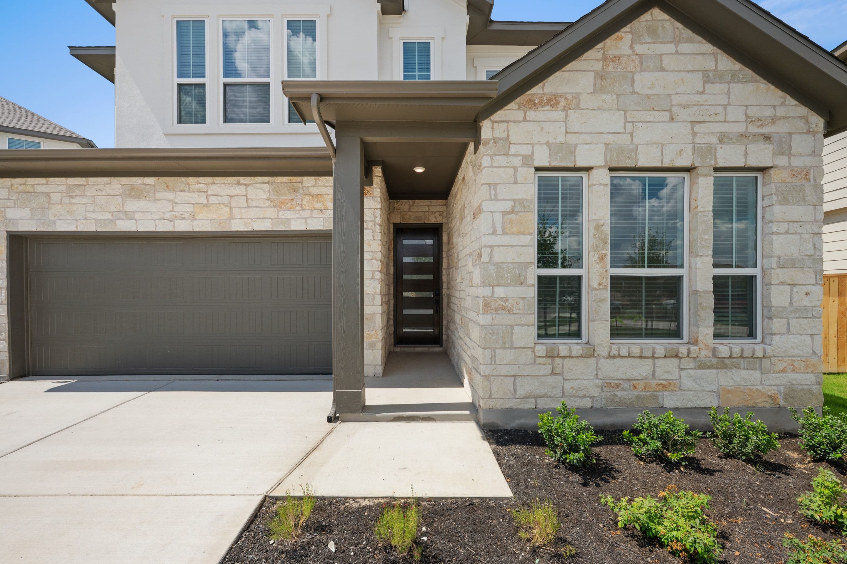 A modern stone and stucco house with a gray garage door and a glass-paneled front door is set against a clear blue sky.