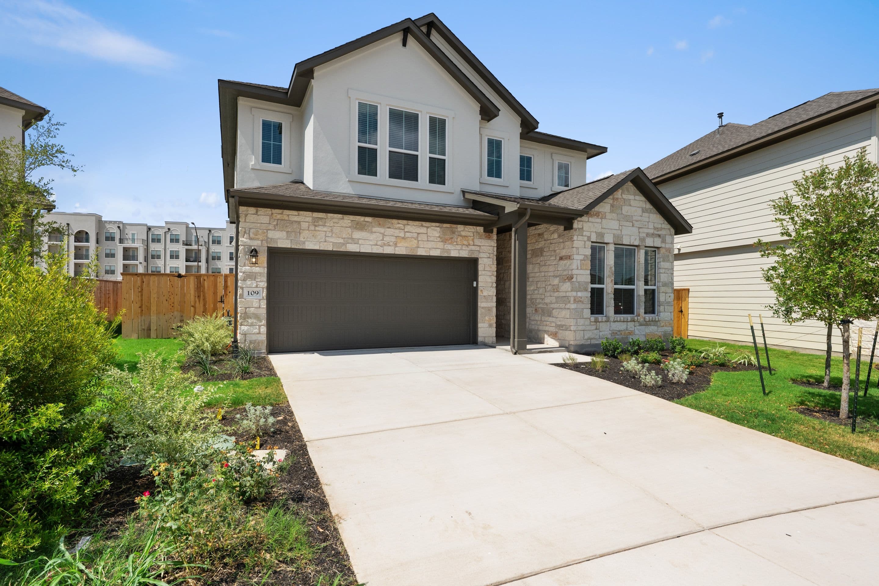 A modern two-story house with a stone and stucco exterior sits on a concrete driveway, with a backdrop of apartment buildings and a clear blue sky.