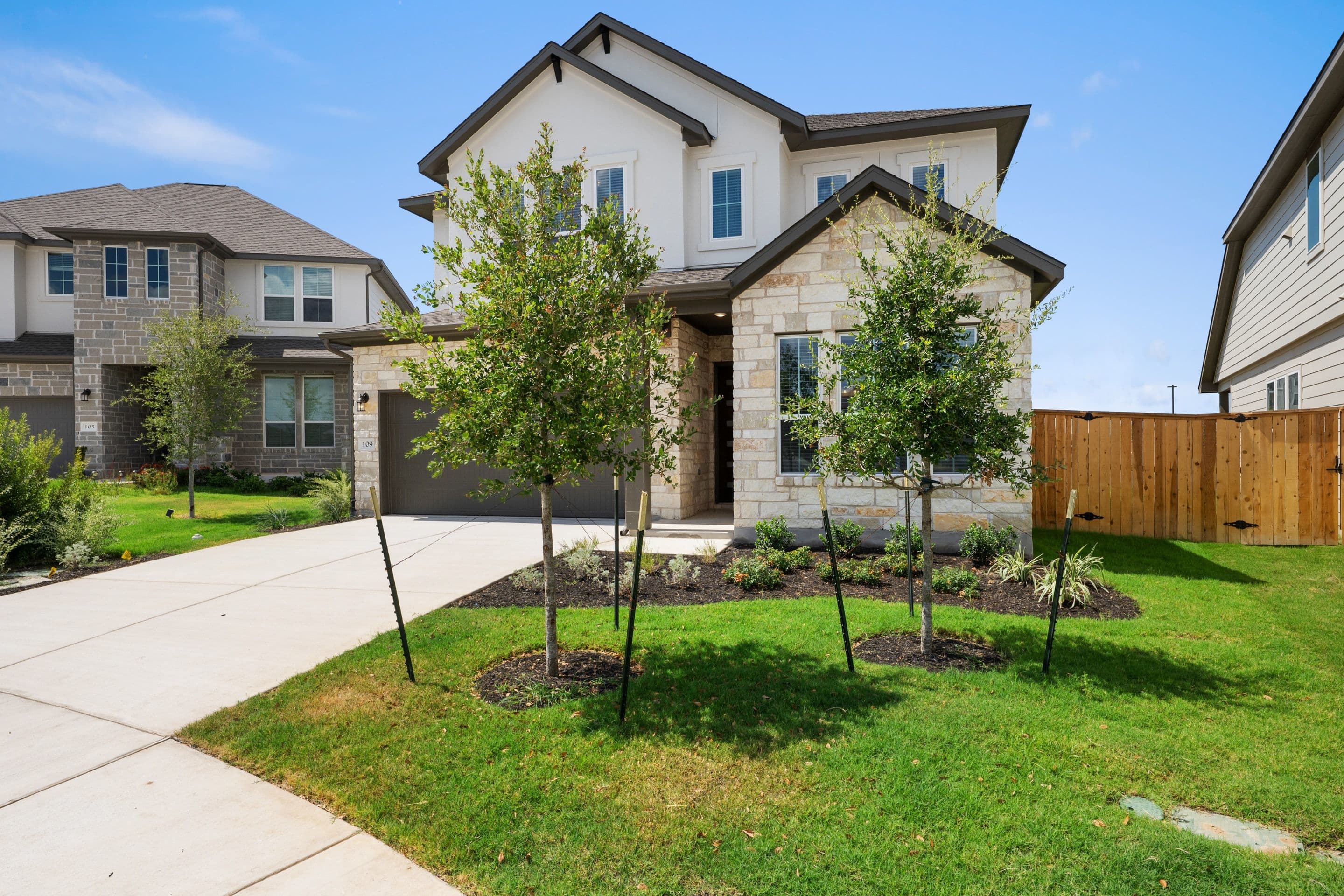 A modern stone and stucco house with a manicured lawn and young trees in the foreground sits under a clear blue sky with neighboring houses visible in the background.