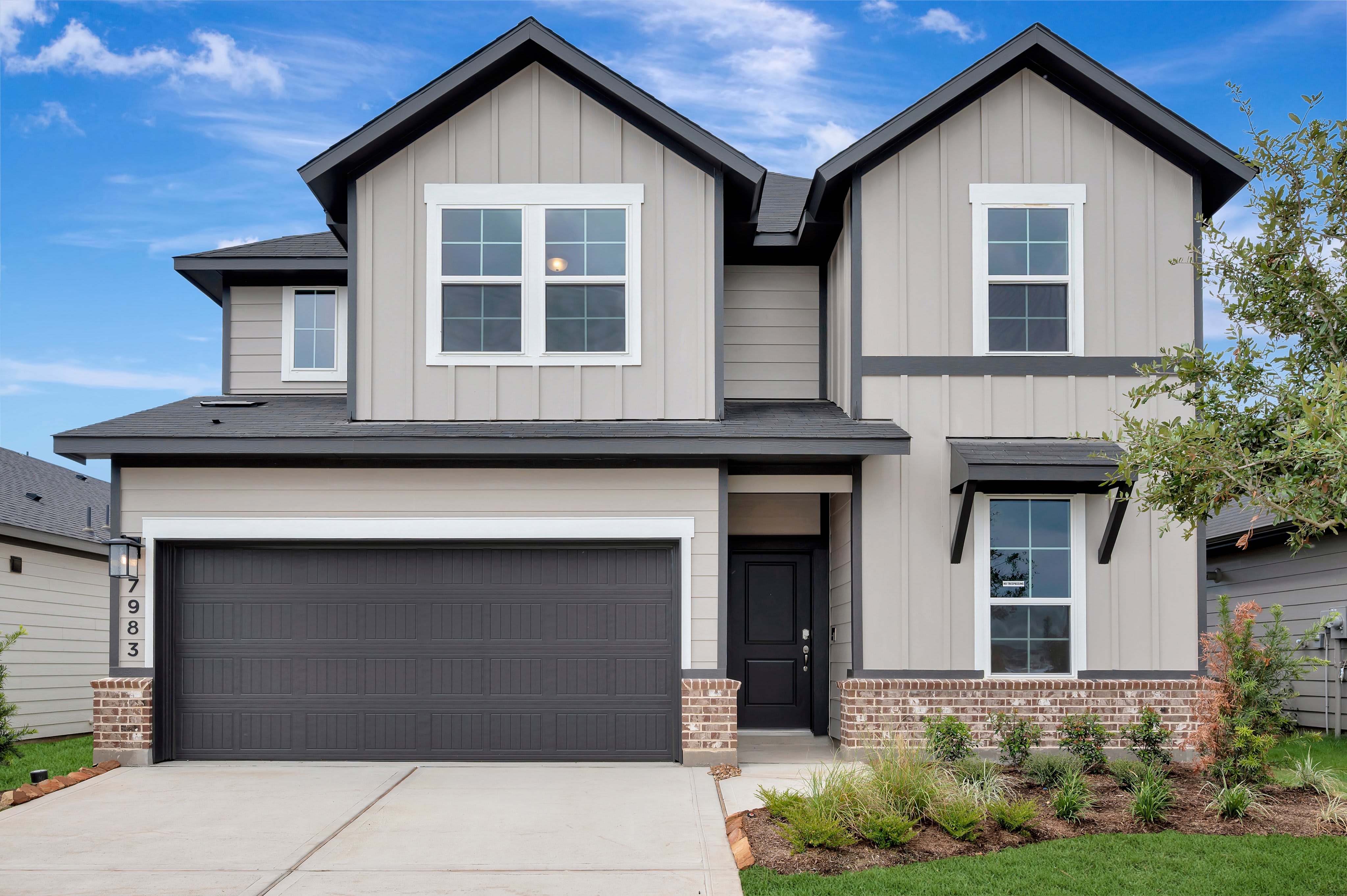 A modern two-story house with a dark garage door and brick accents in the foreground stands against a bright blue sky with scattered clouds.