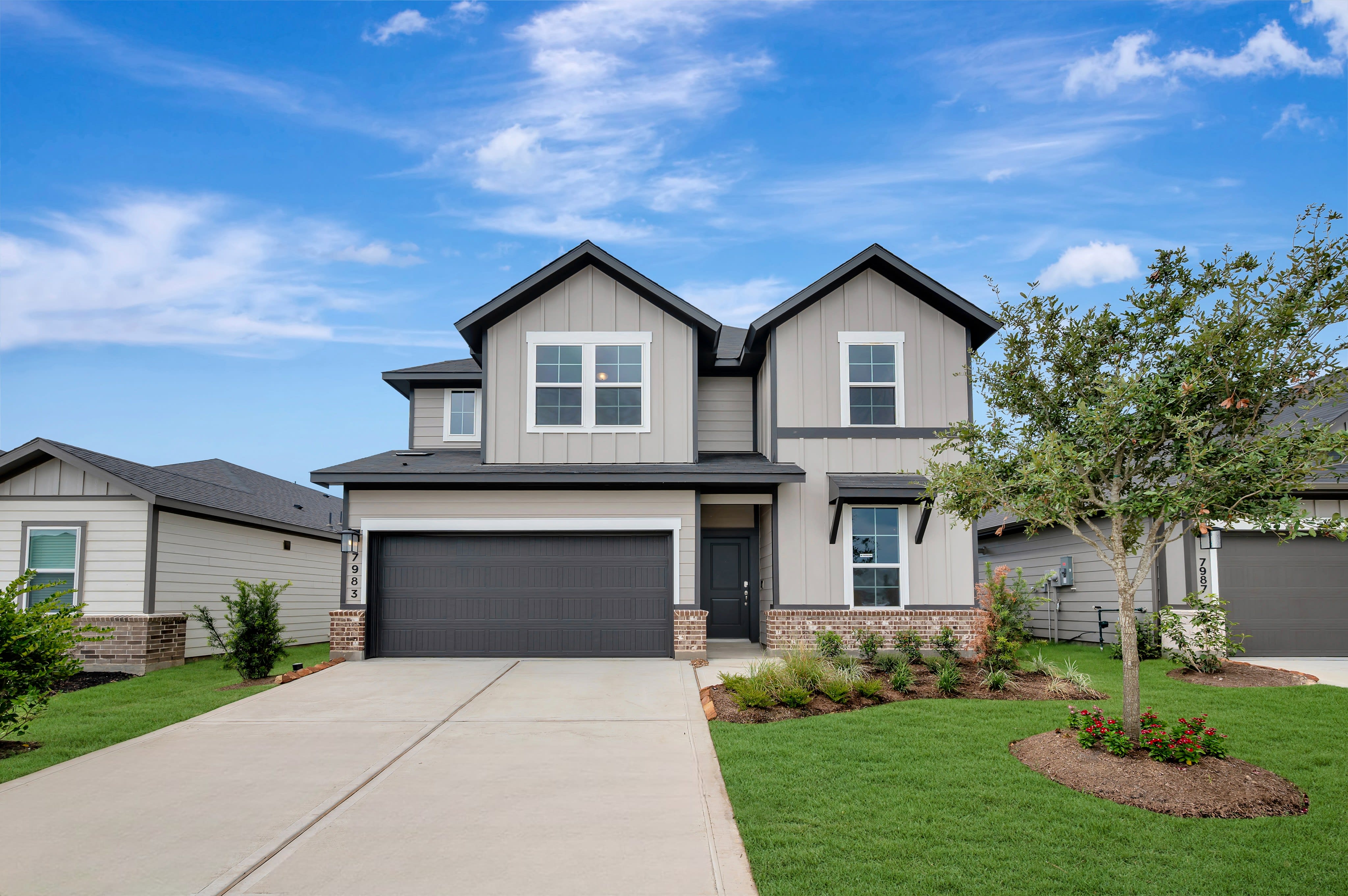 A modern two-story house with a gray garage door and manicured lawn sits under a bright blue sky with scattered clouds.
