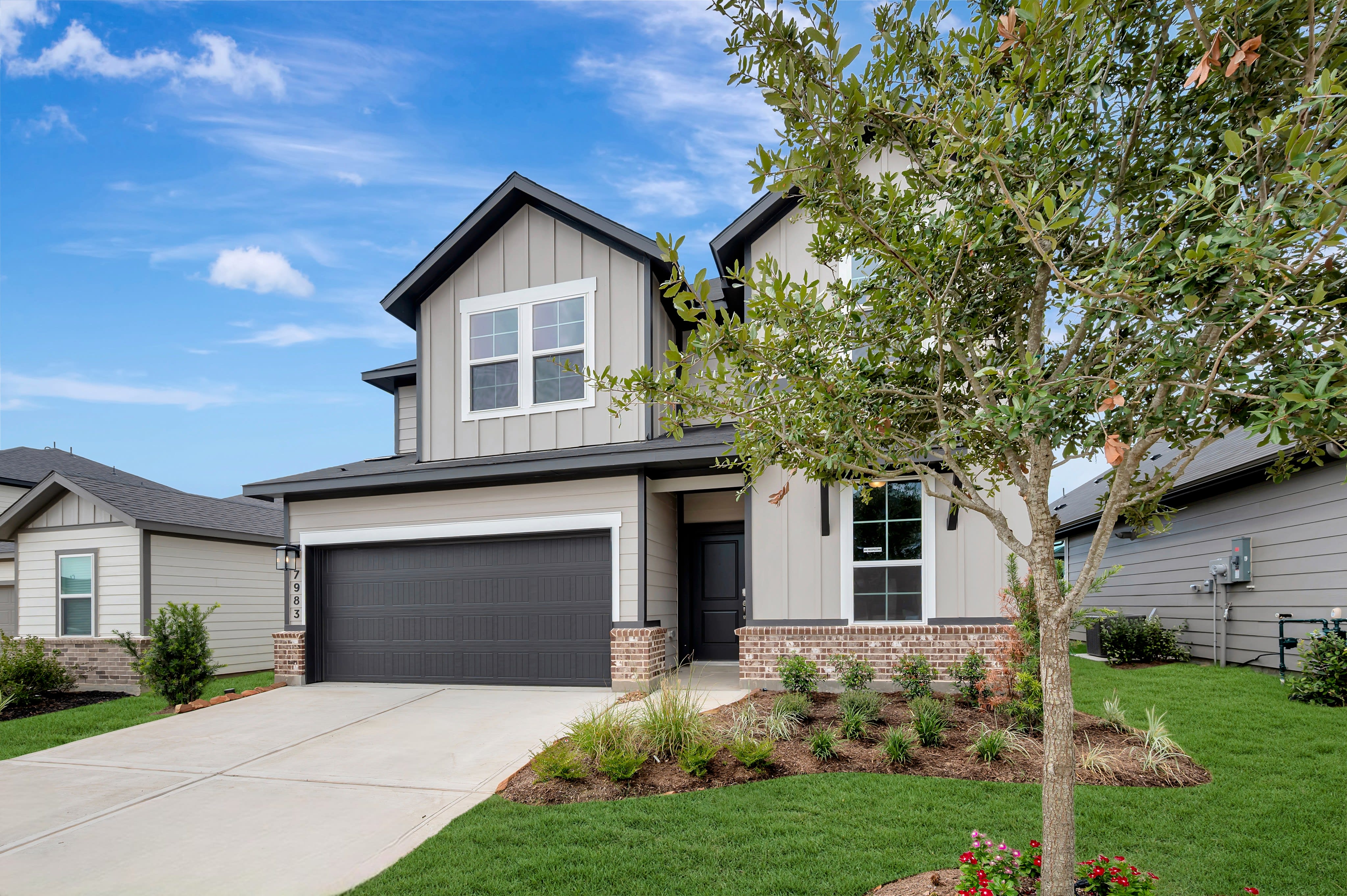A modern gray house with a dark garage door sits on a manicured lawn in front of a bright blue sky with scattered clouds.