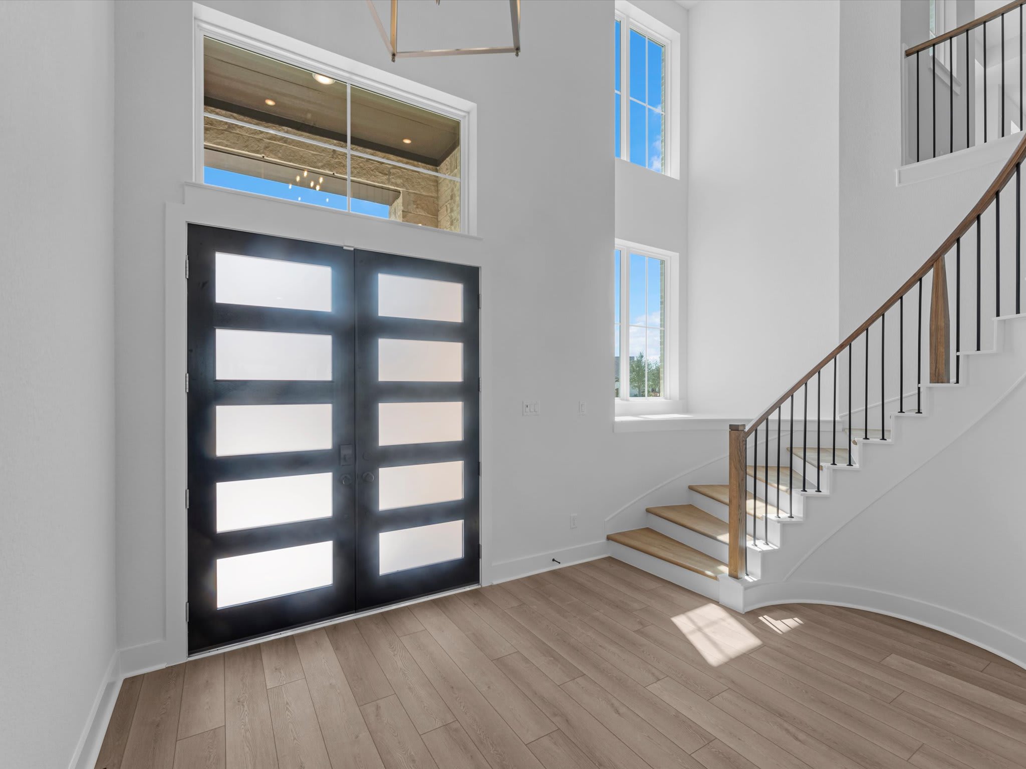 A modern entryway with a frosted glass door and light wood flooring in the foreground, and a grand staircase with a view of the outdoors through a large window in the background.