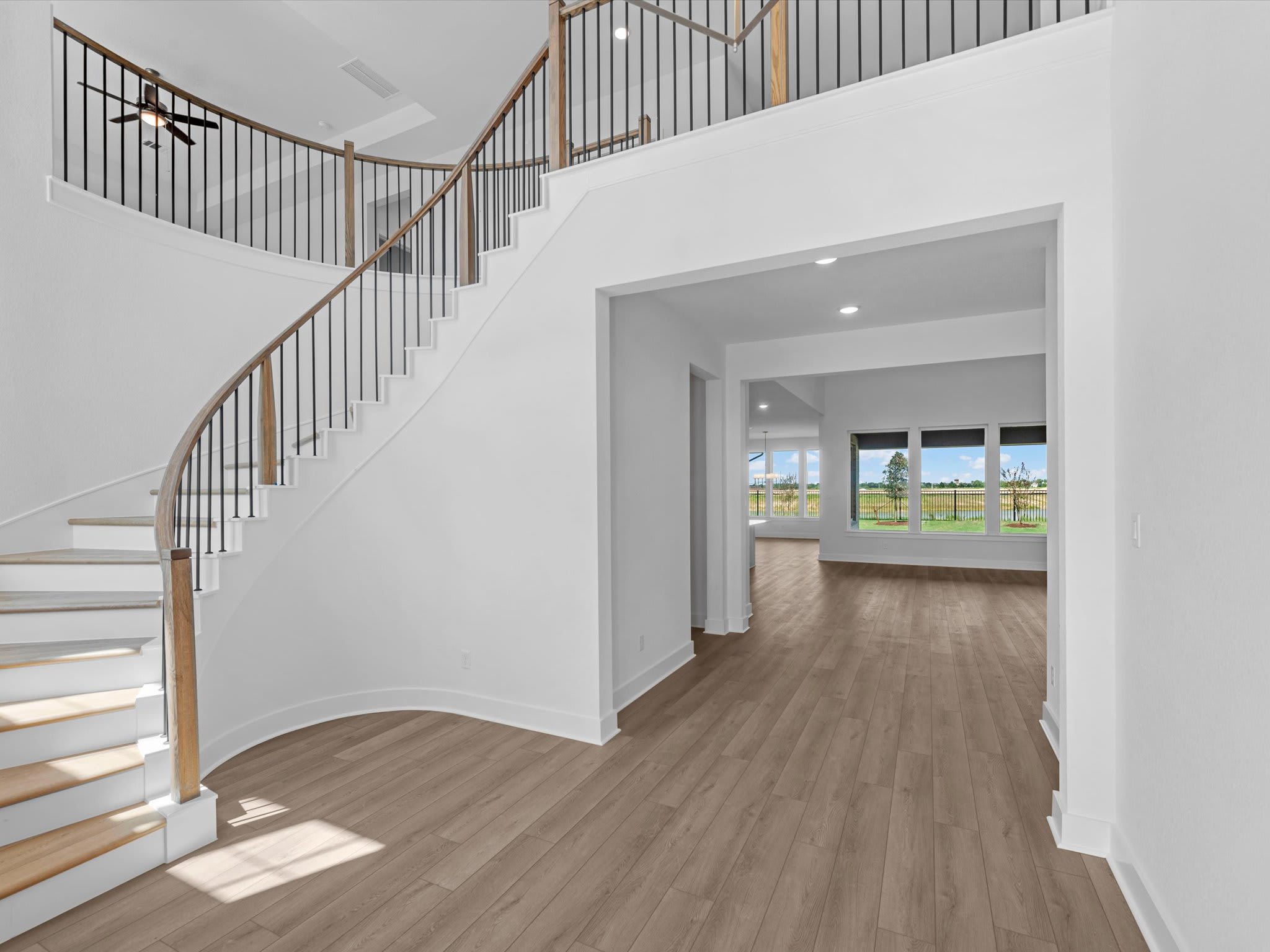 A grand curved staircase with wooden banisters dominates the foreground, leading to a bright, open living space with large windows overlooking a green field in the background.