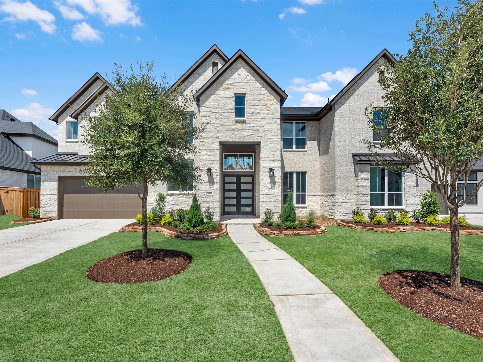 A modern stone house with a manicured lawn and trees in the foreground is set against a bright blue sky with scattered clouds in the background.