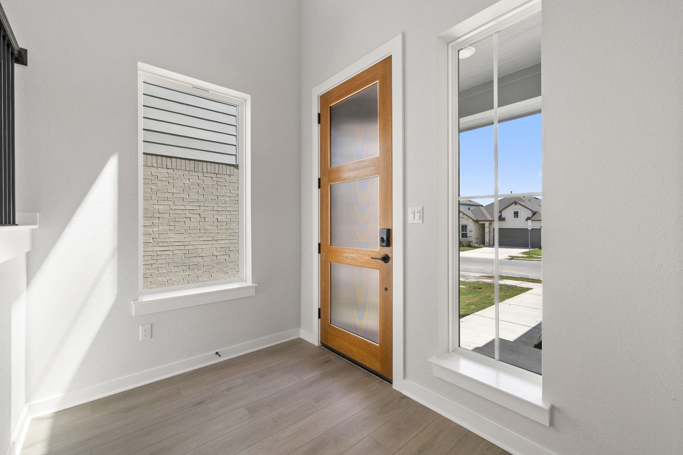 A modern wooden front door with frosted glass panels is centered in the foreground, flanked by tall windows, while the background reveals a suburban street with houses and a clear blue sky.