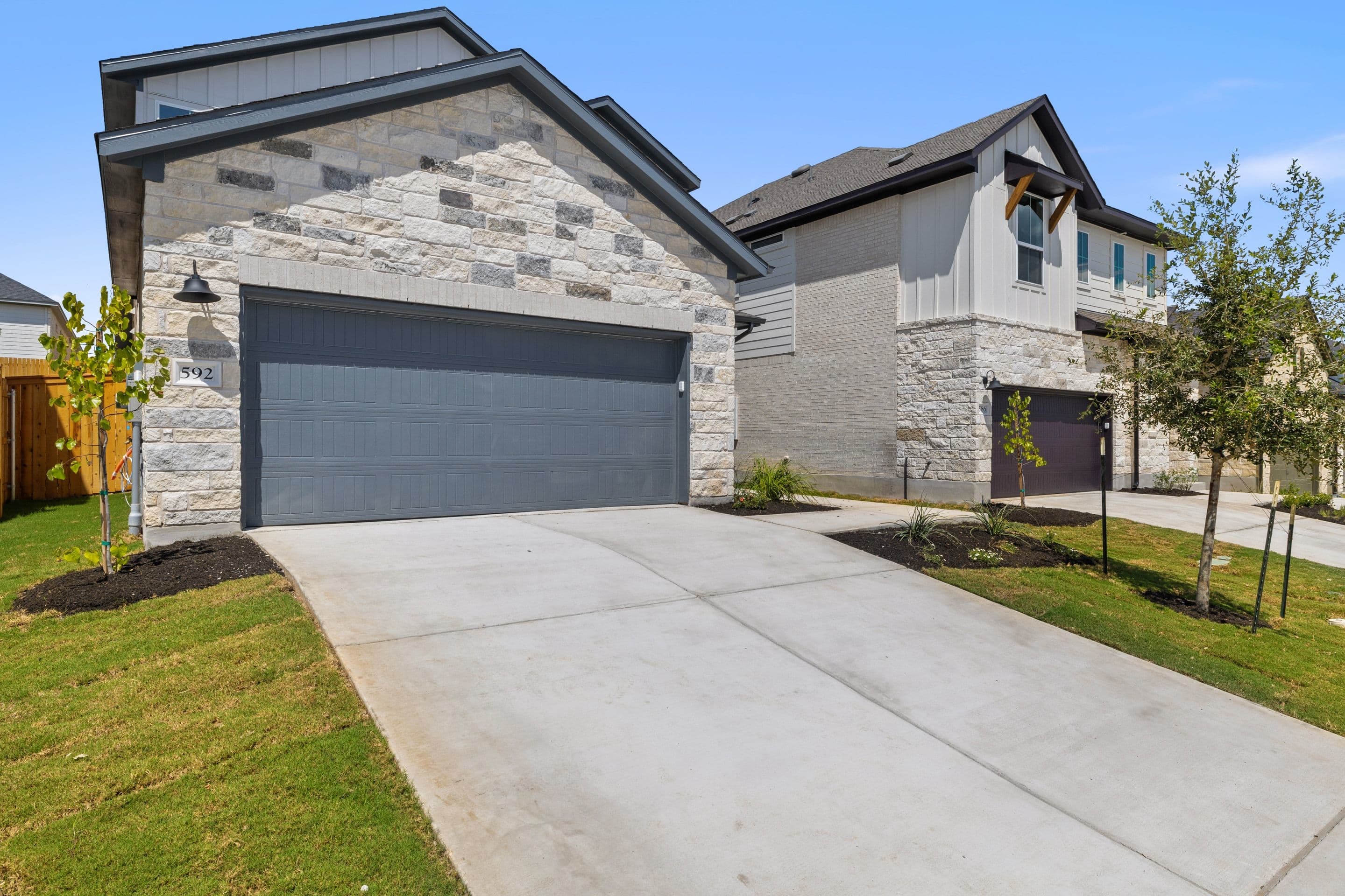 A concrete driveway leads to a modern house with a stone facade in the foreground, while another similar house and trees are visible in the background under a clear blue sky.