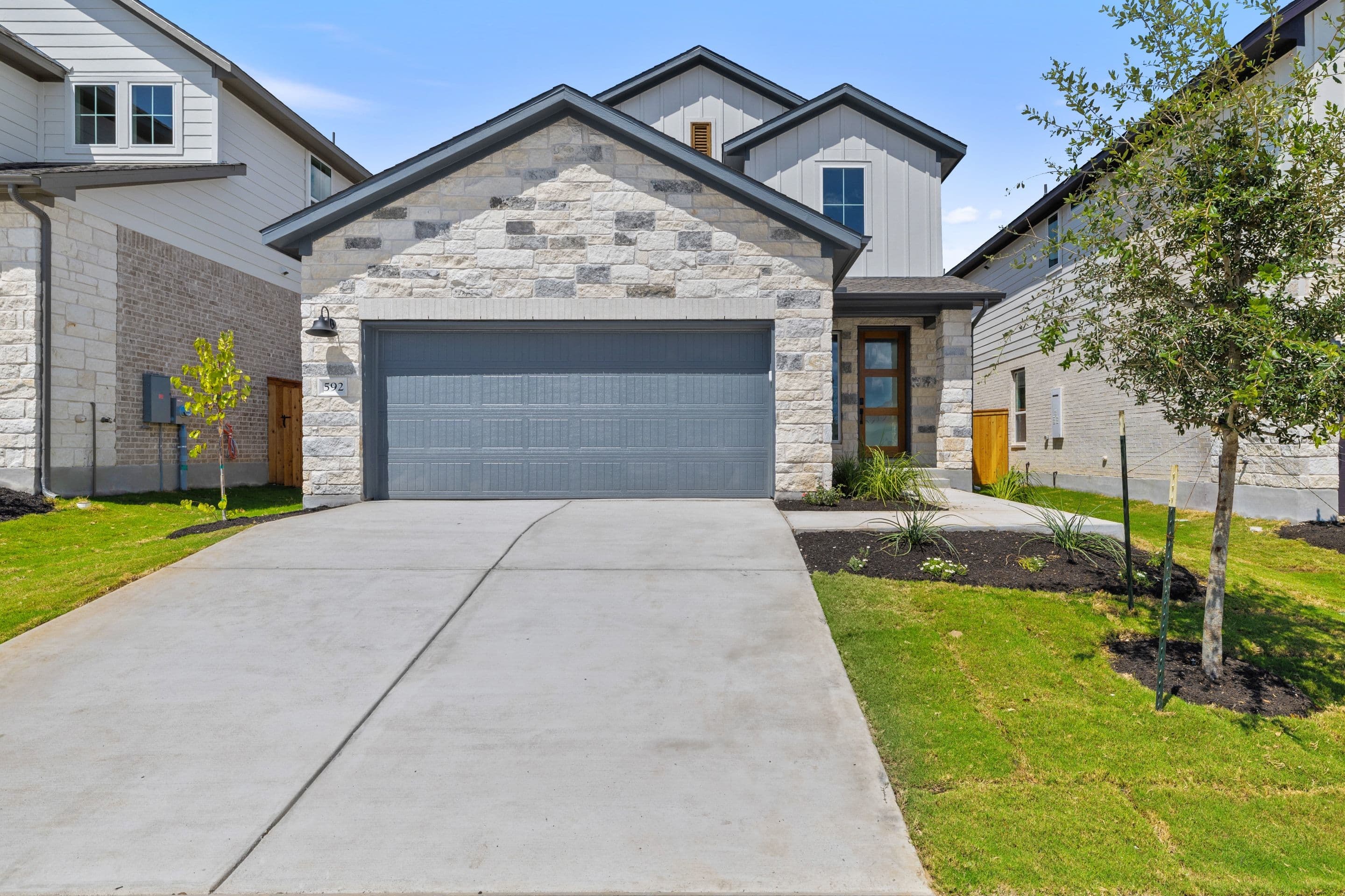 A modern stone-clad house with a gray garage door sits on a concrete driveway, with manicured lawns and trees in the foreground and neighboring houses in the background.
