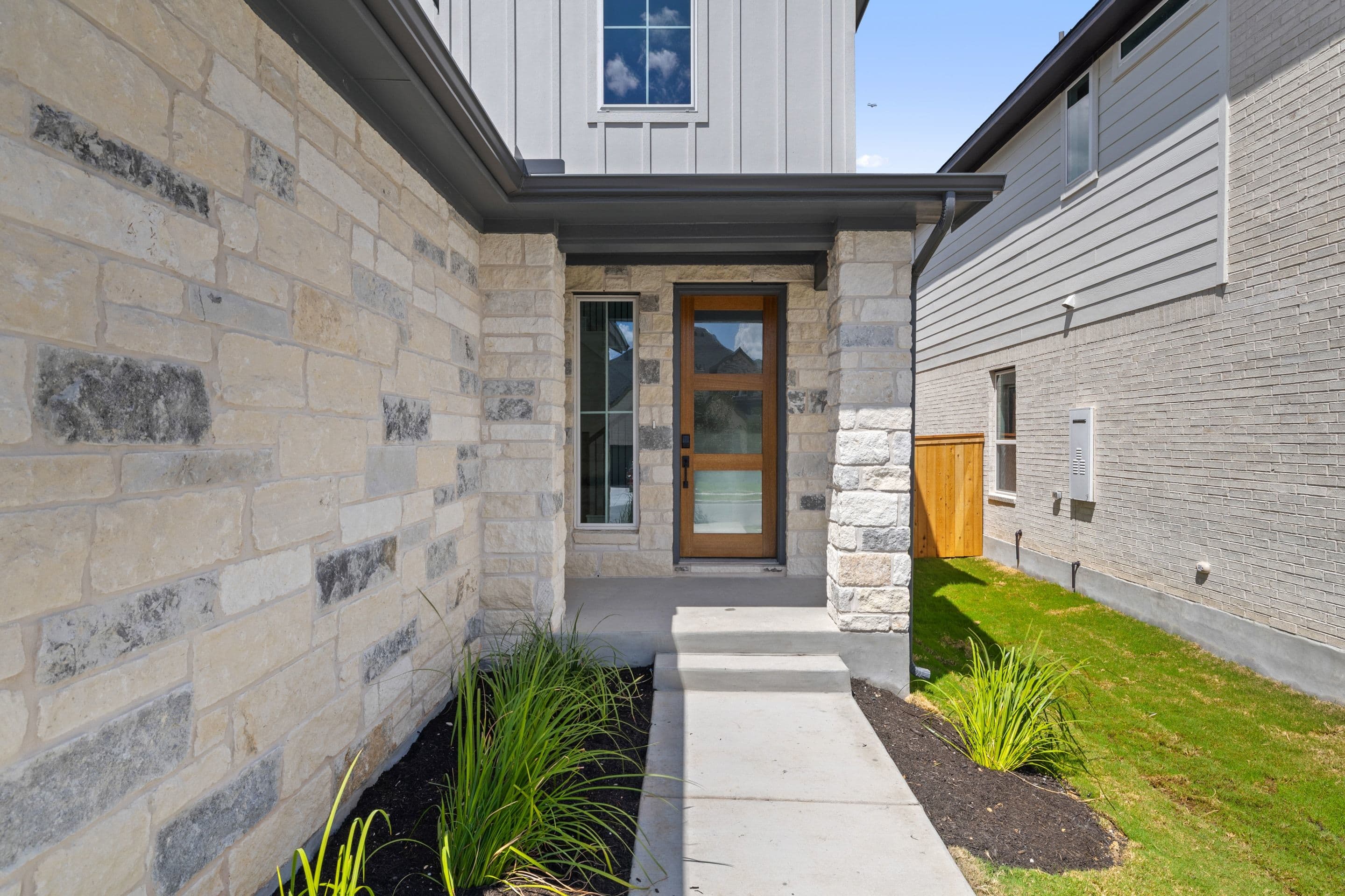 A modern home with a stone facade and a wooden door is framed by lush green grass and landscaping.