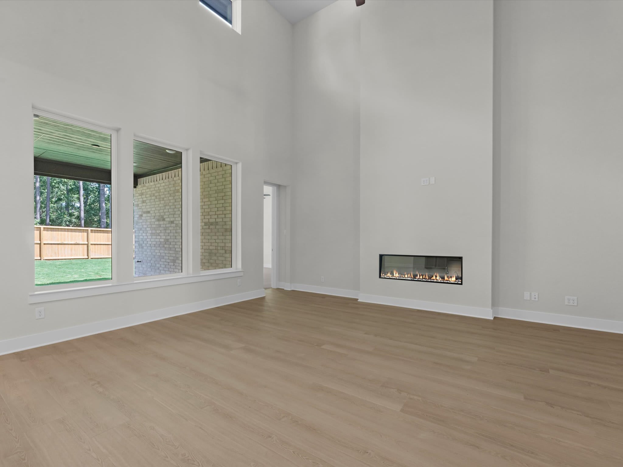 A modern living room with a light wood floor in the foreground and a sleek fireplace on the right wall, while the background features large windows overlooking a backyard with a fence and trees.