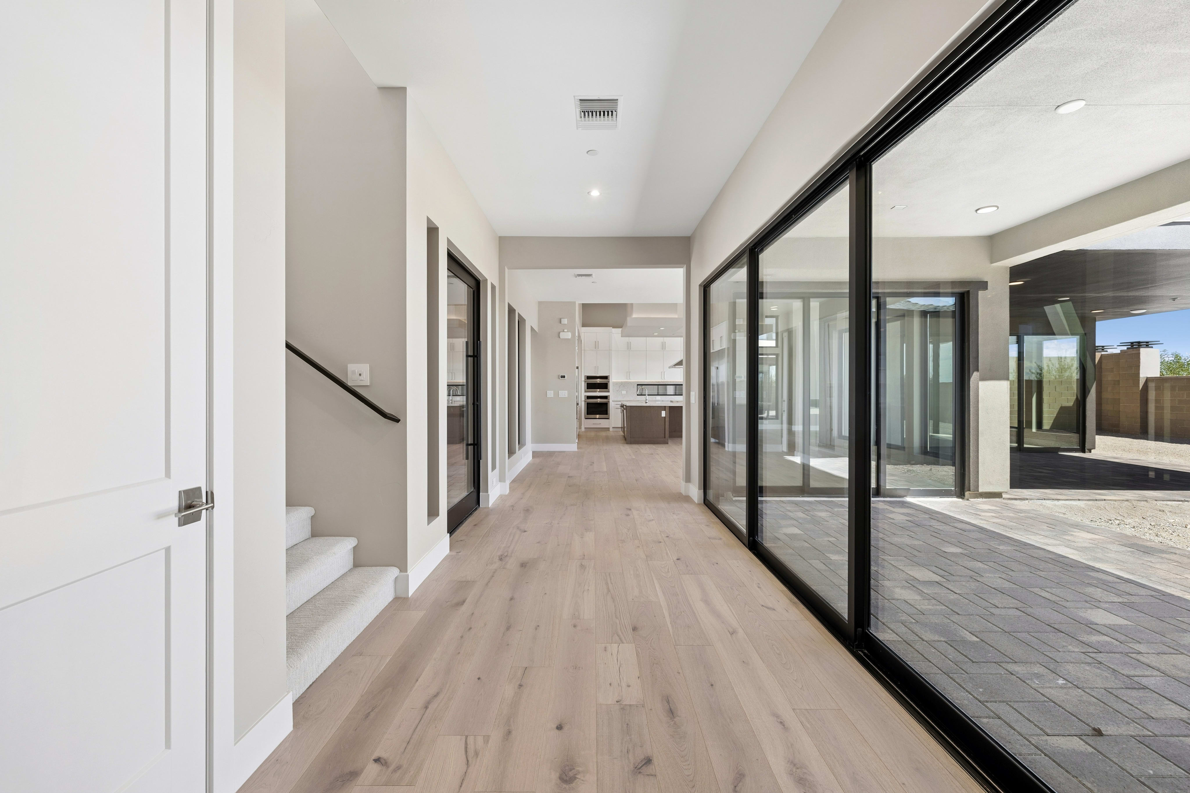 A modern hallway with a light wood floor and white doors in the foreground opens to a bright kitchen and an outdoor patio with large sliding glass doors in the background.