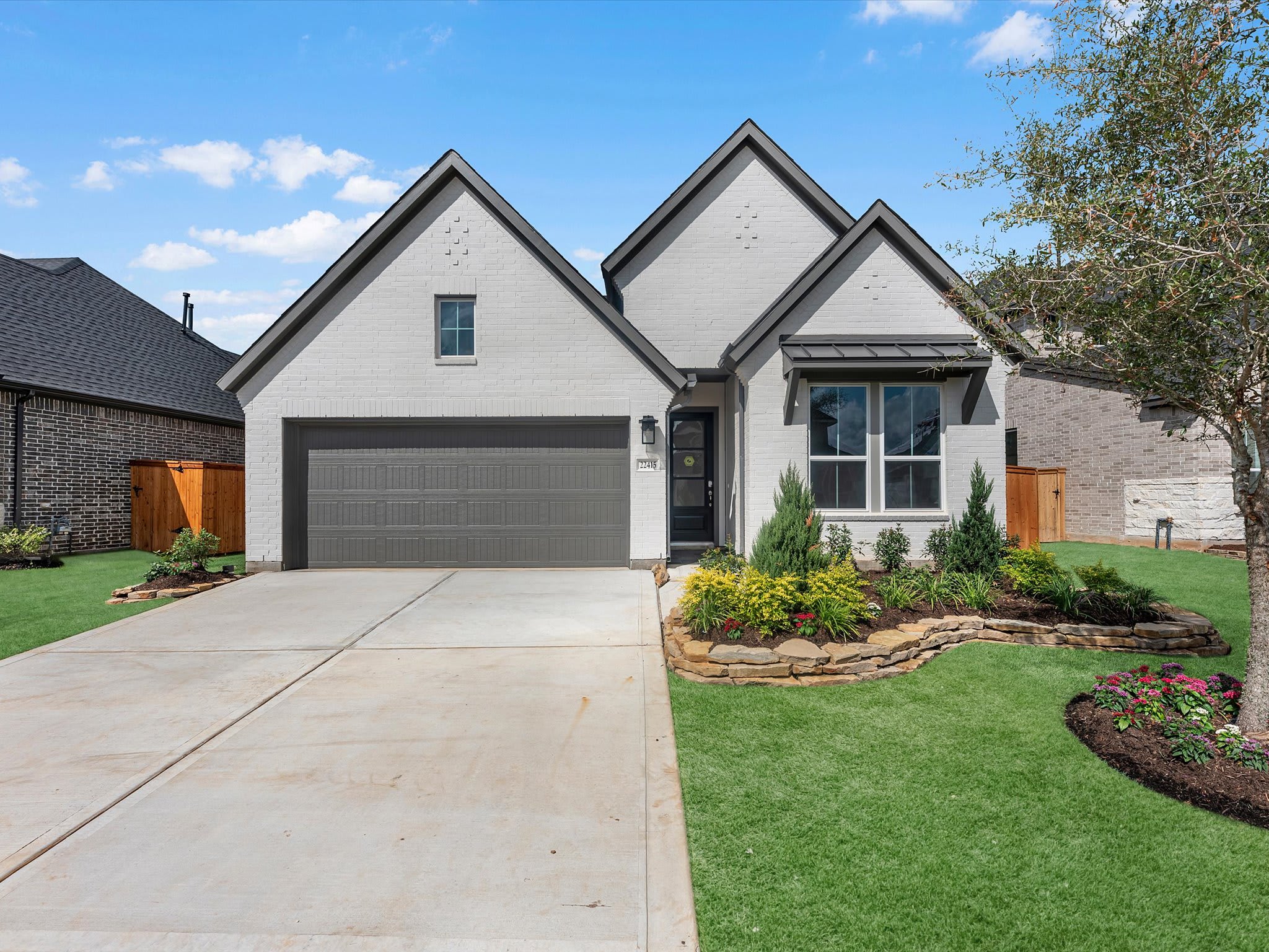 A modern white brick house with a gray garage door and manicured landscaping sits on a concrete driveway, with neighboring houses and a blue sky with clouds in the background.