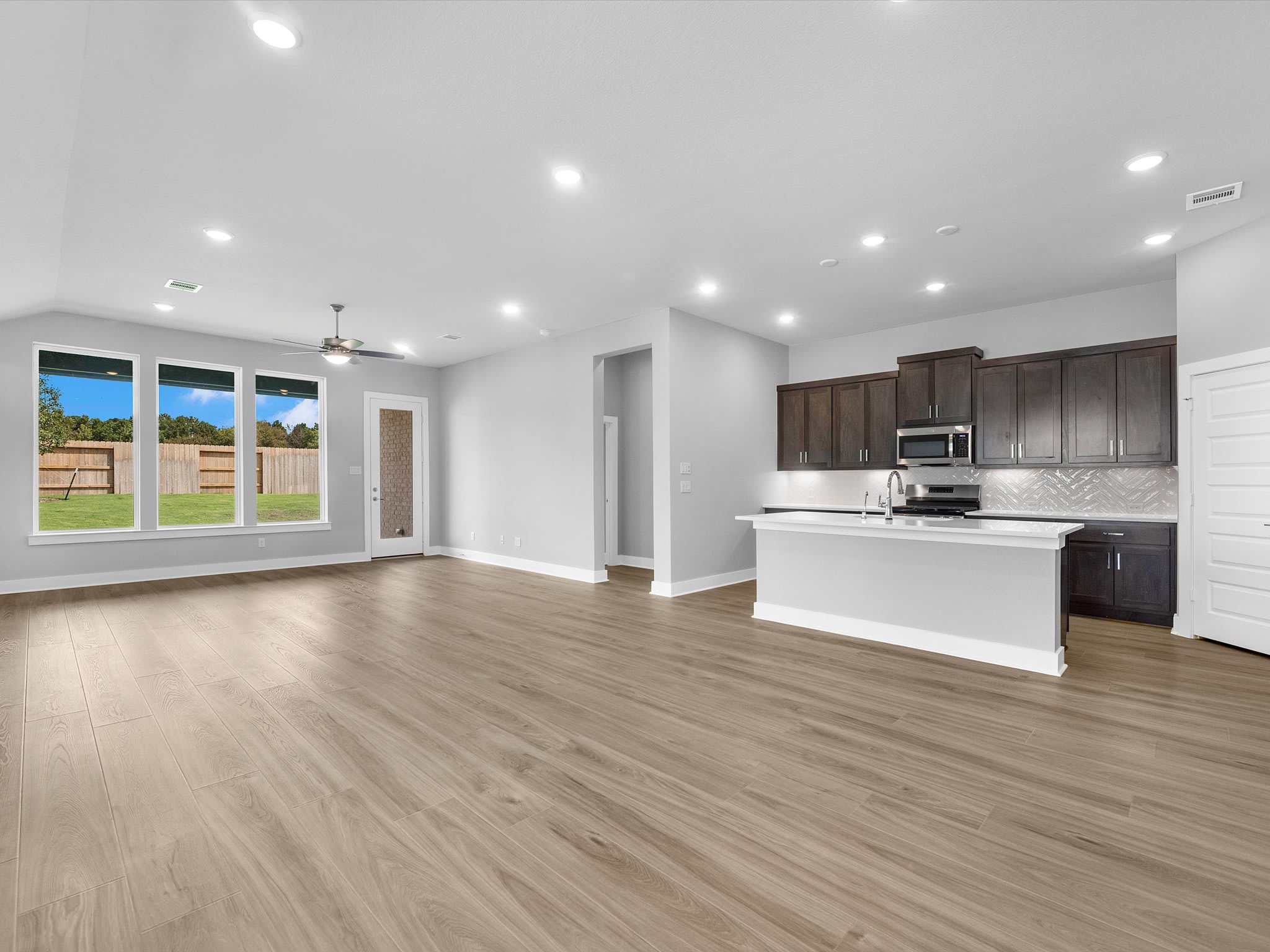 A spacious, modern living area with light wood flooring in the foreground opens to a kitchen with dark cabinets and a white island in the background, beyond which are large windows overlooking a green lawn and blue sky.