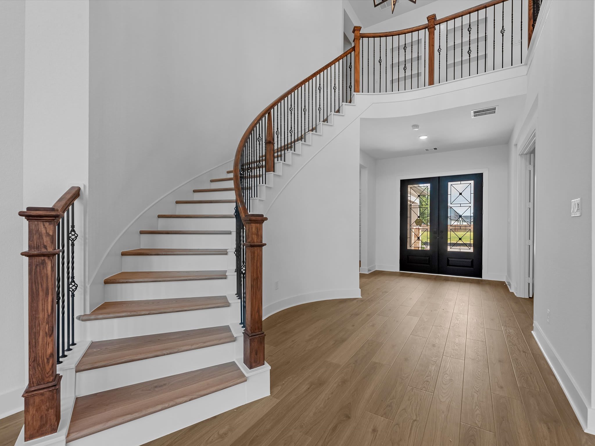 A curved wooden staircase with black iron railings leads upward from a light-colored hardwood floor toward a second-story balcony and a black double front door in the background.