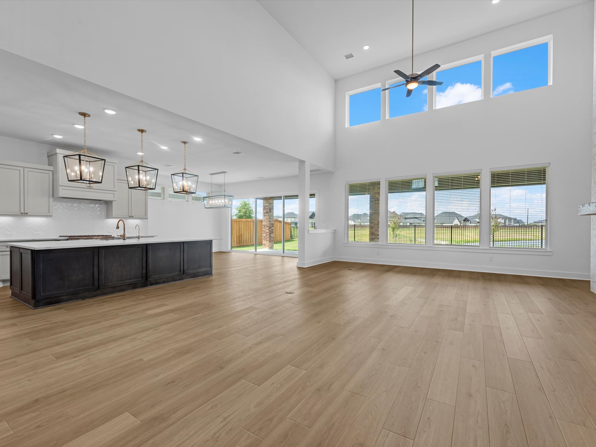 A modern open-concept living area features light wood flooring and a dark kitchen island in the foreground, with large windows and a high ceiling in the background.