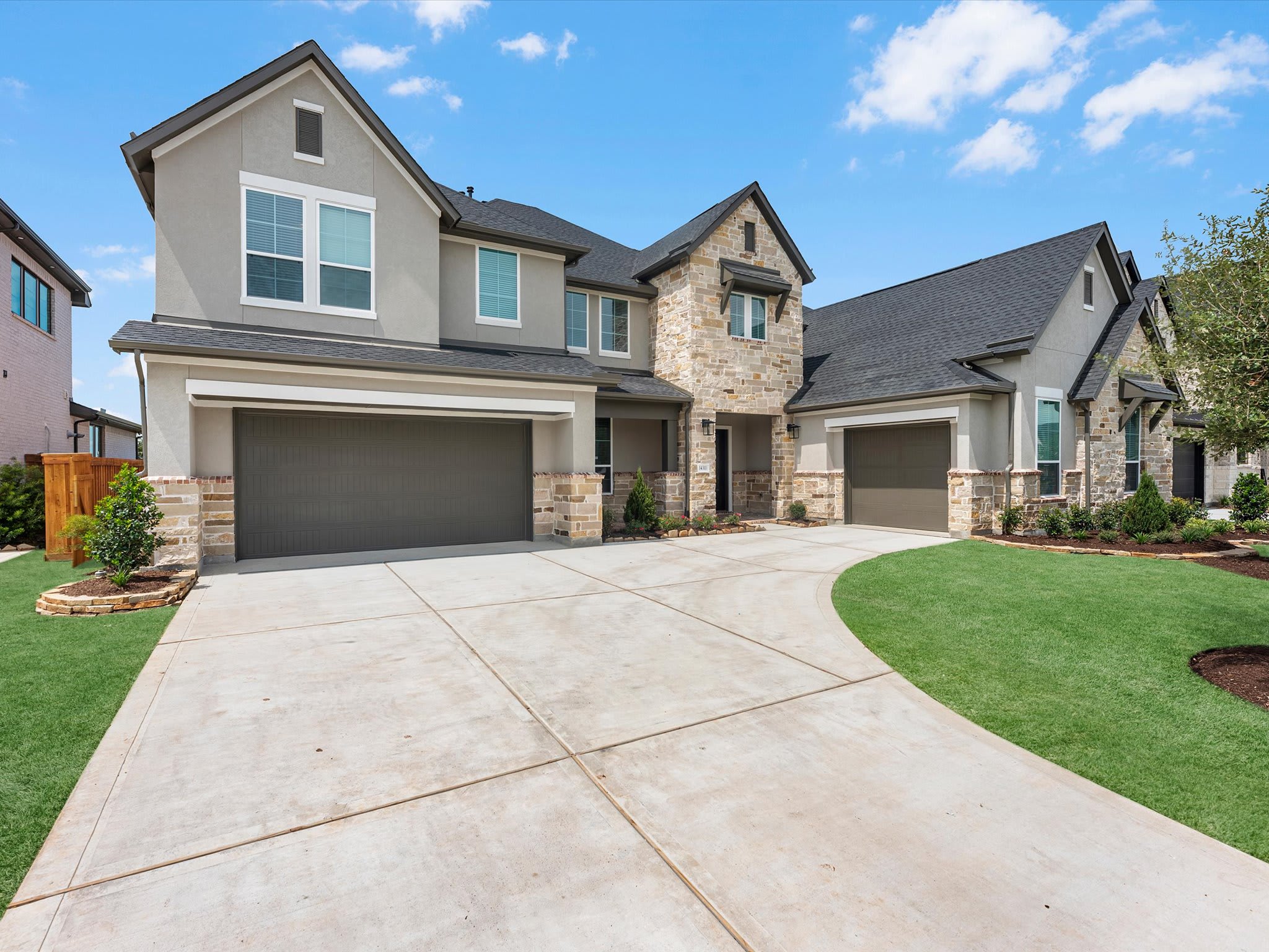 A wide concrete driveway leads toward a modern two-story home with grey siding and stone accents under a bright blue sky with scattered clouds.
