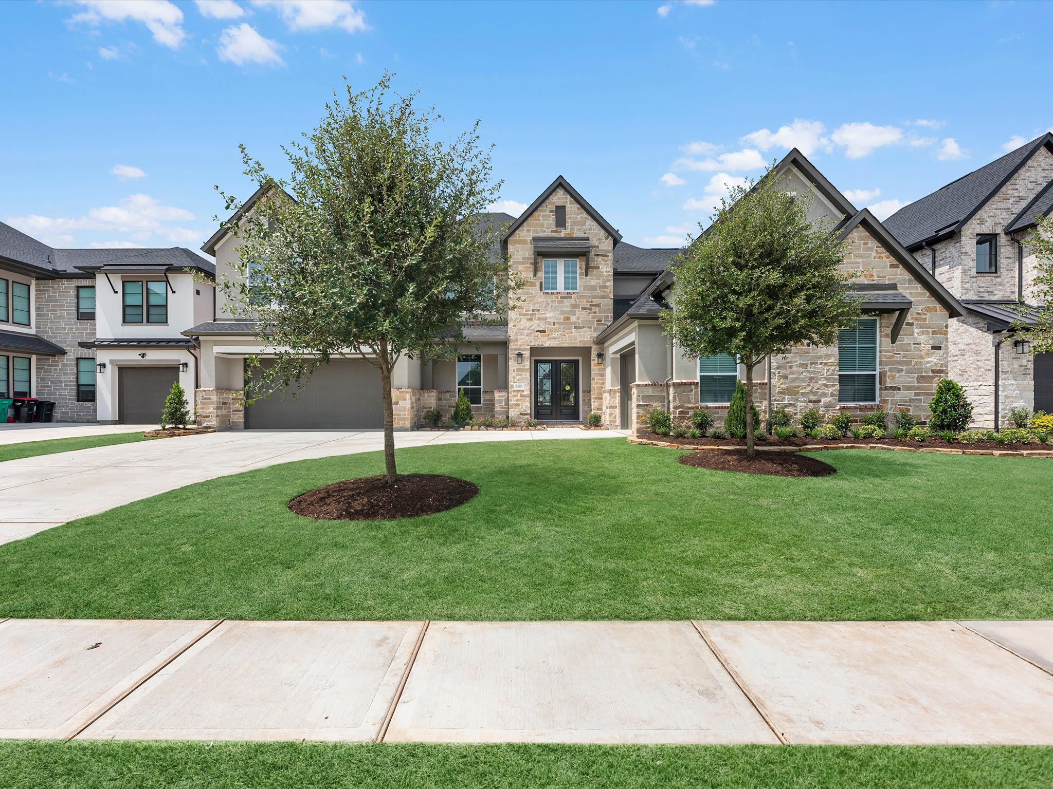 A manicured green lawn and concrete sidewalk lead toward a large, modern stone and siding house under a bright blue sky.