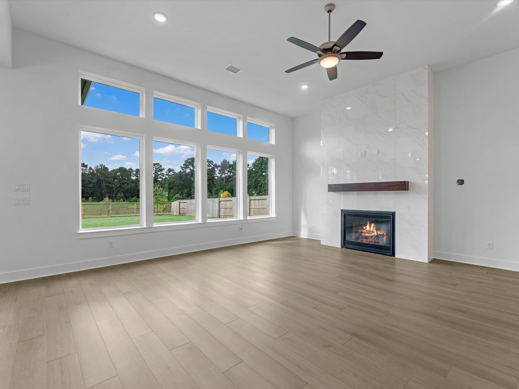 An empty room with light wood floors and a marble fireplace in the foreground, with large windows overlooking a green yard in the background.