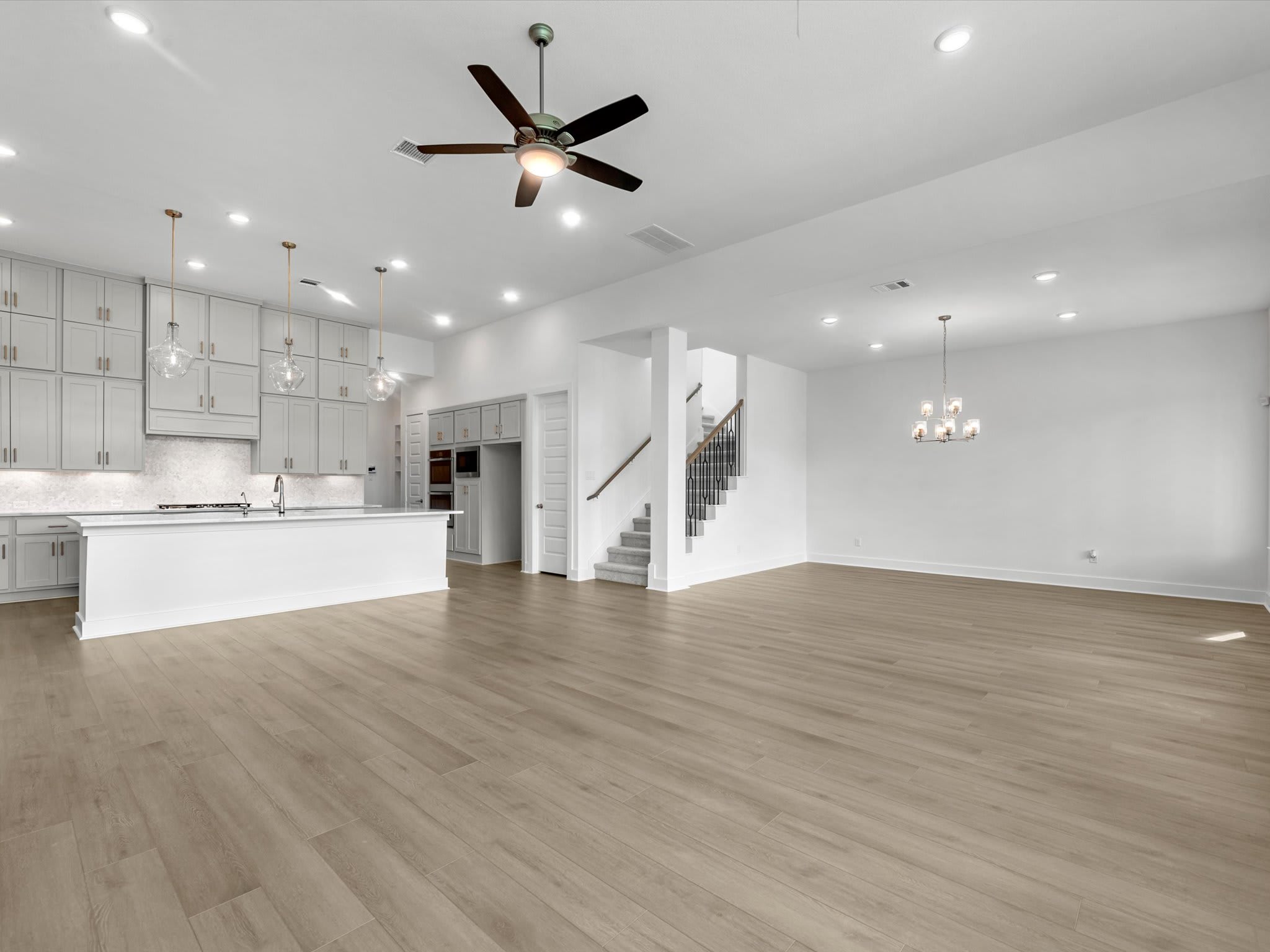 A spacious open-concept living area features light wood flooring in the foreground, with a white kitchen and a staircase leading upstairs in the background.
