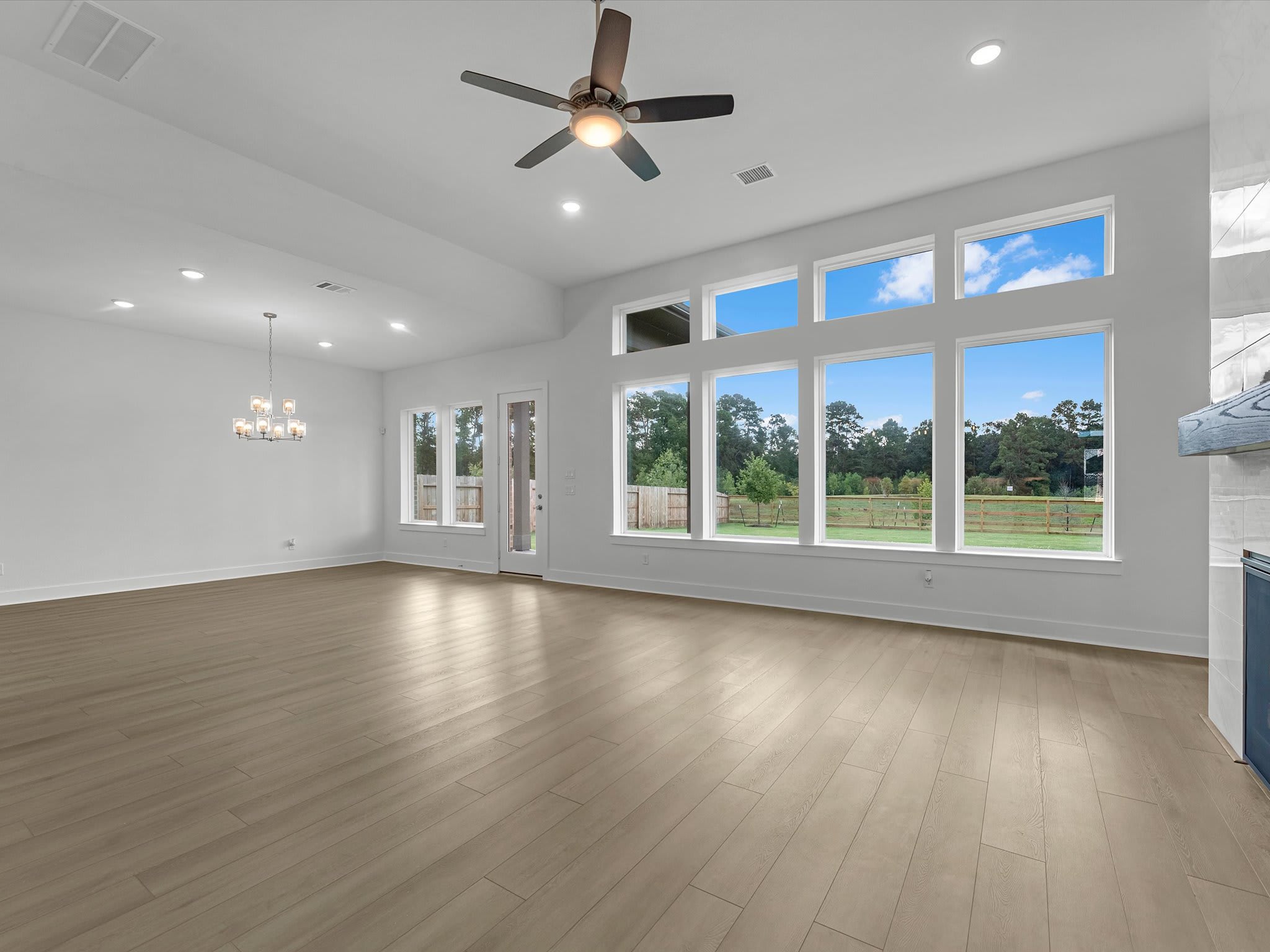 An empty room with light wood flooring is illuminated by a ceiling fan and large windows overlooking a green landscape and blue sky.
