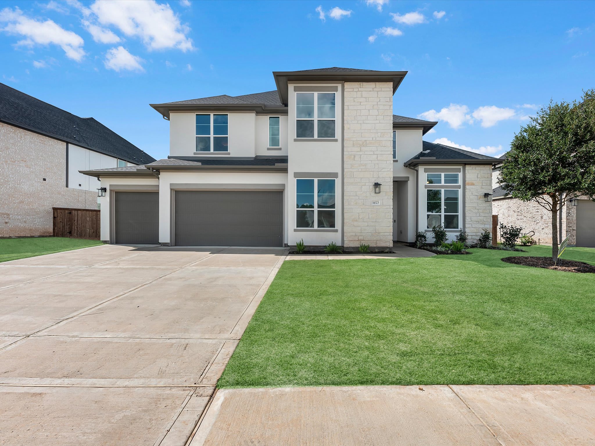 A modern two-story house with a concrete driveway and green lawn sits under a bright blue sky with scattered clouds.