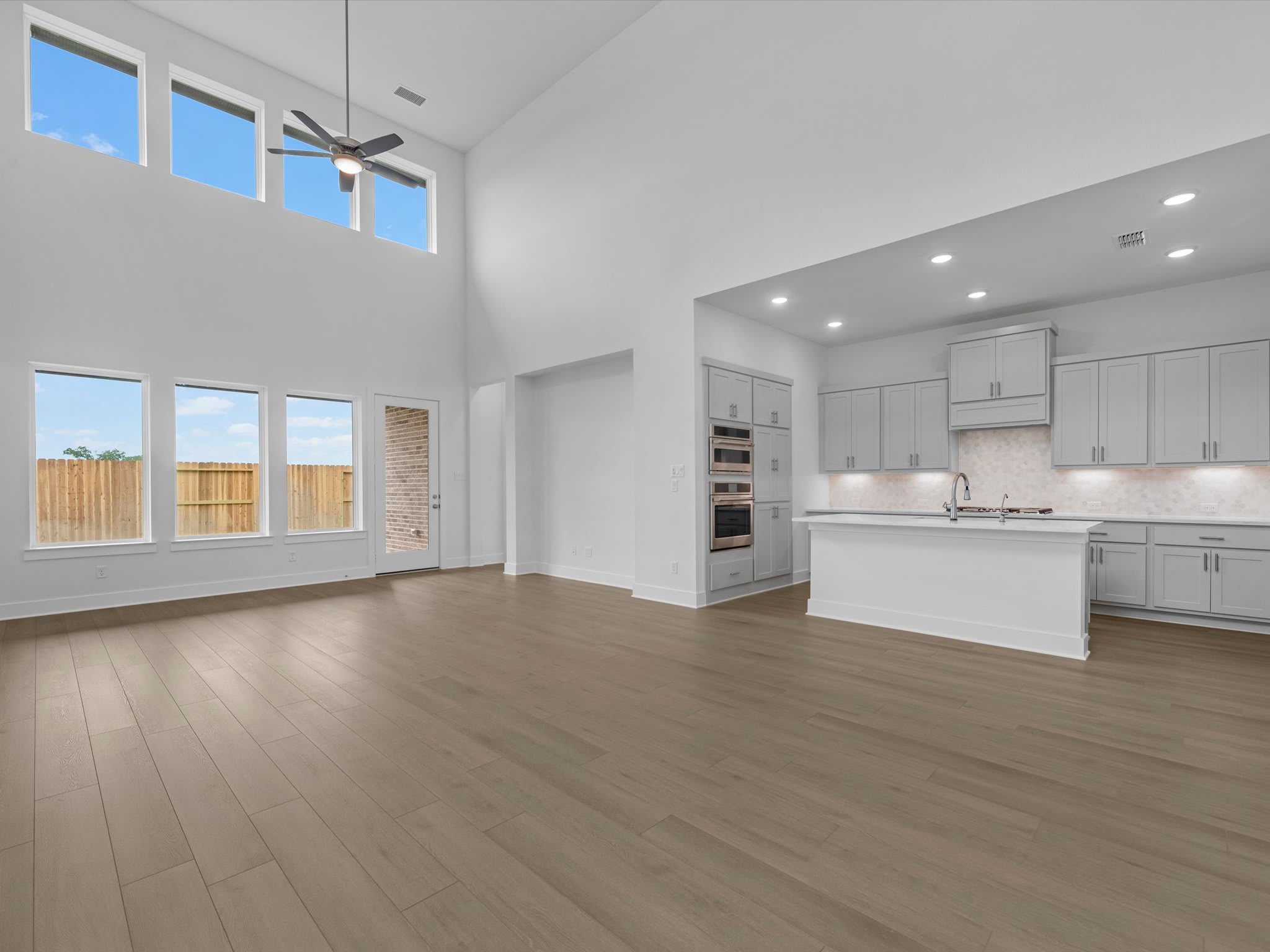 An open-concept living area with light wood flooring in the foreground leads to a modern white kitchen and high ceilings with large windows in the background.