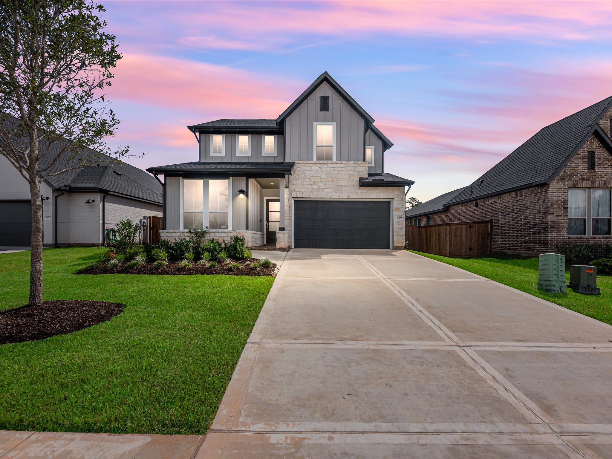 A concrete driveway and green lawn lead to a modern grey and stone house under a colorful sunset sky.