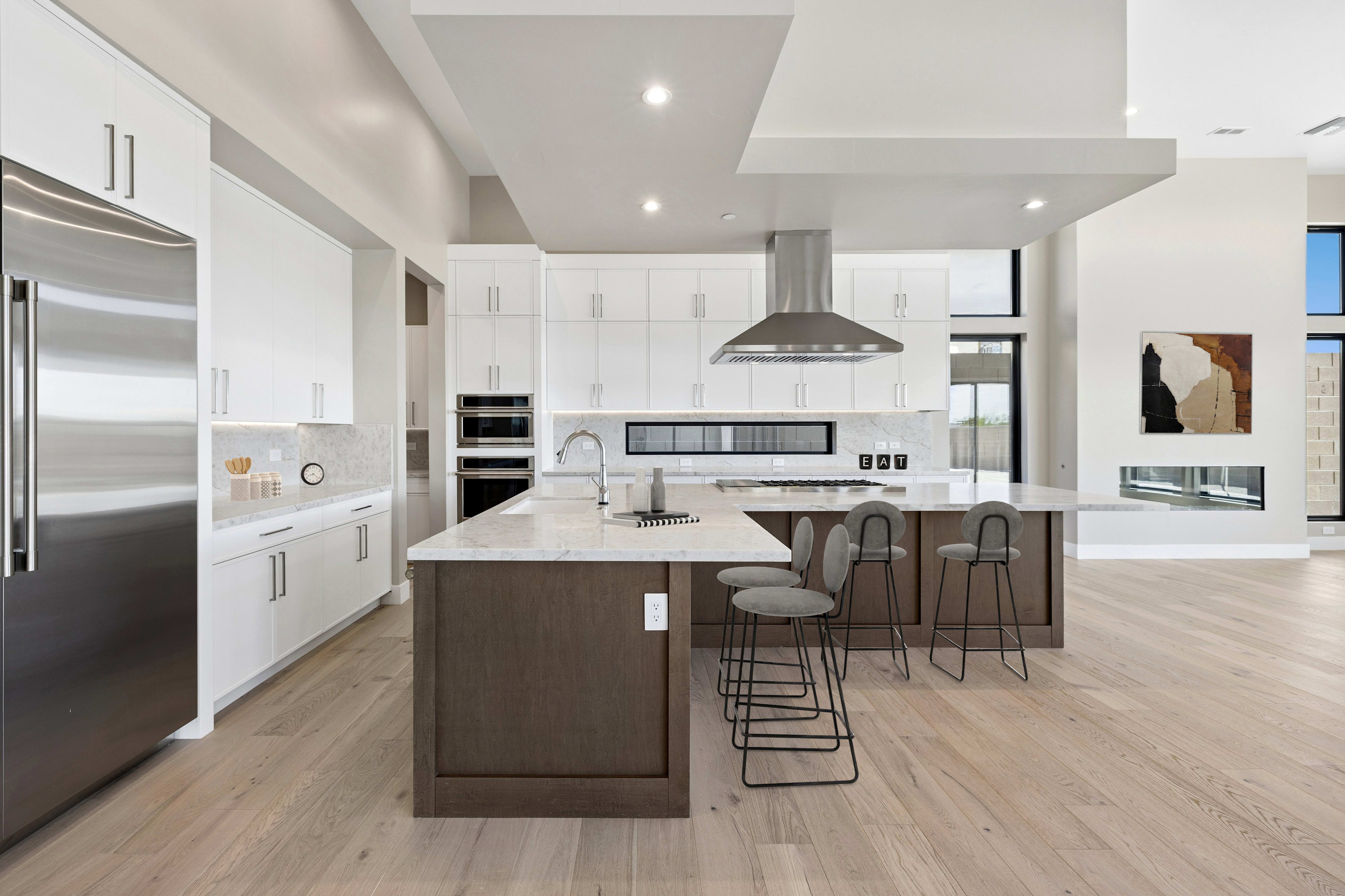 A modern kitchen features a large dark wood island with grey barstools in the foreground and white cabinetry with stainless steel appliances in the background.
