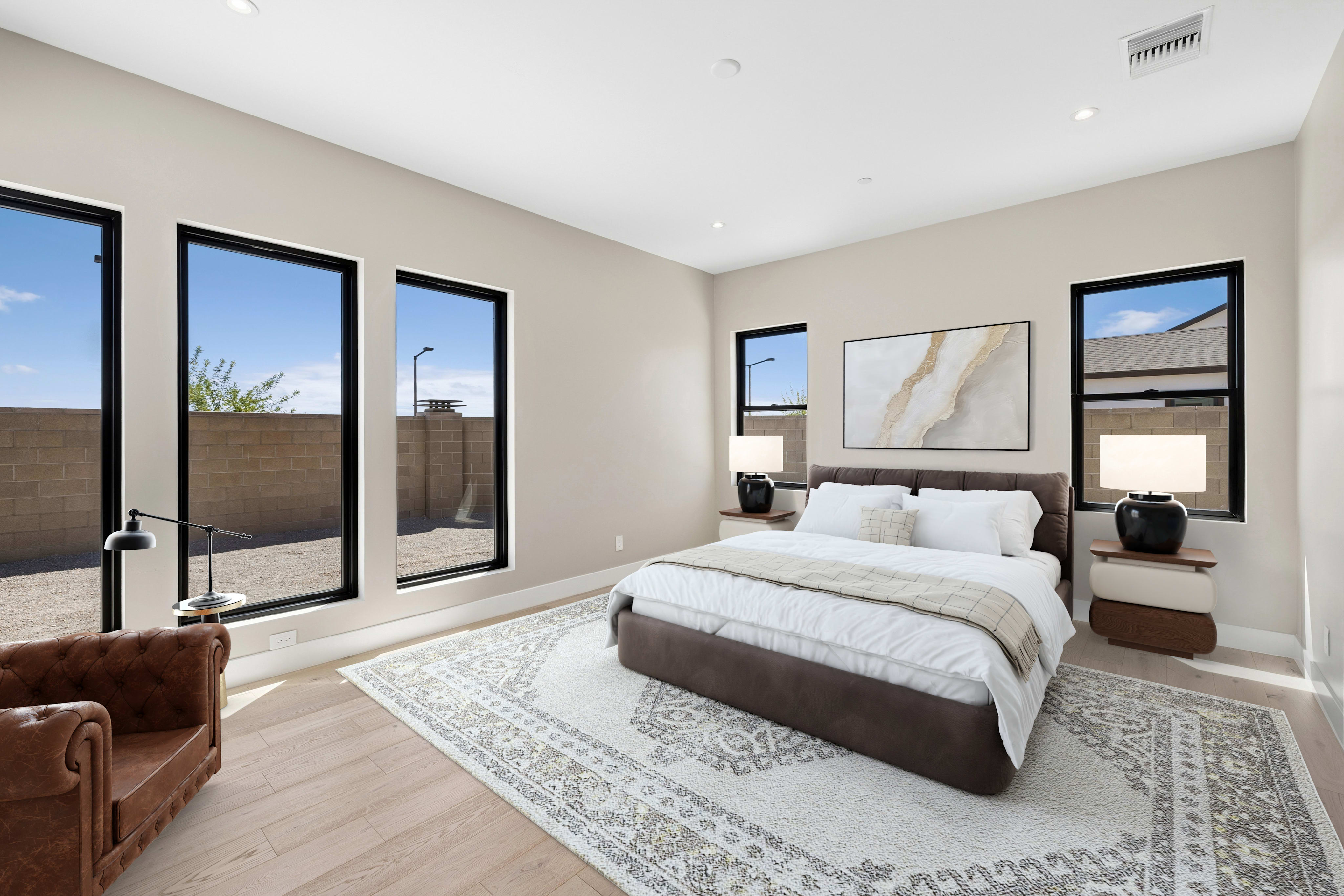 A brown leather armchair sits in the foreground of a modern bedroom featuring a large bed on a patterned rug and several black-framed windows looking out onto a walled courtyard.