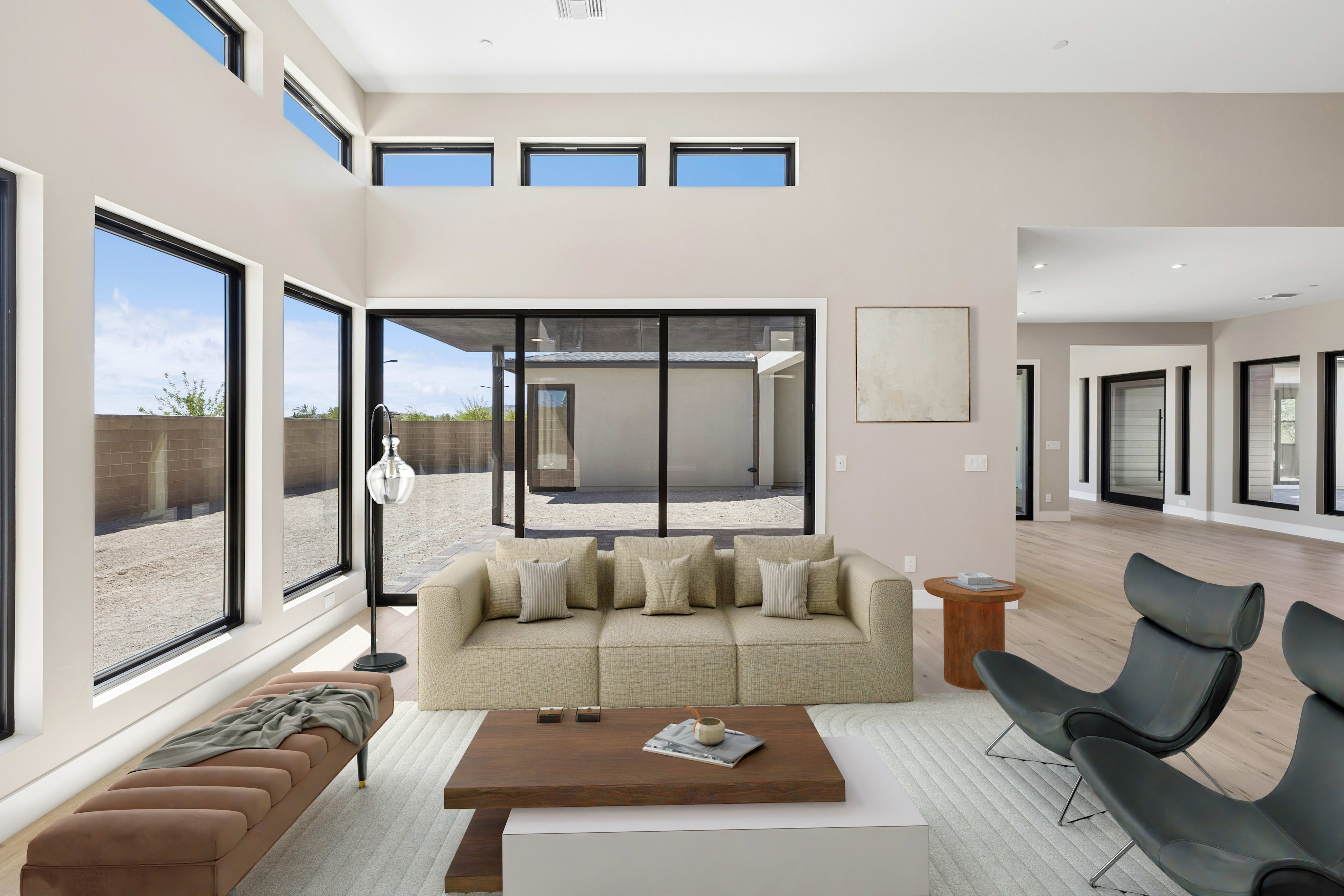 A modern living room with a beige sofa, grey lounge chairs, and a wooden coffee table sits before large windows and sliding glass doors that look out onto a desert courtyard.