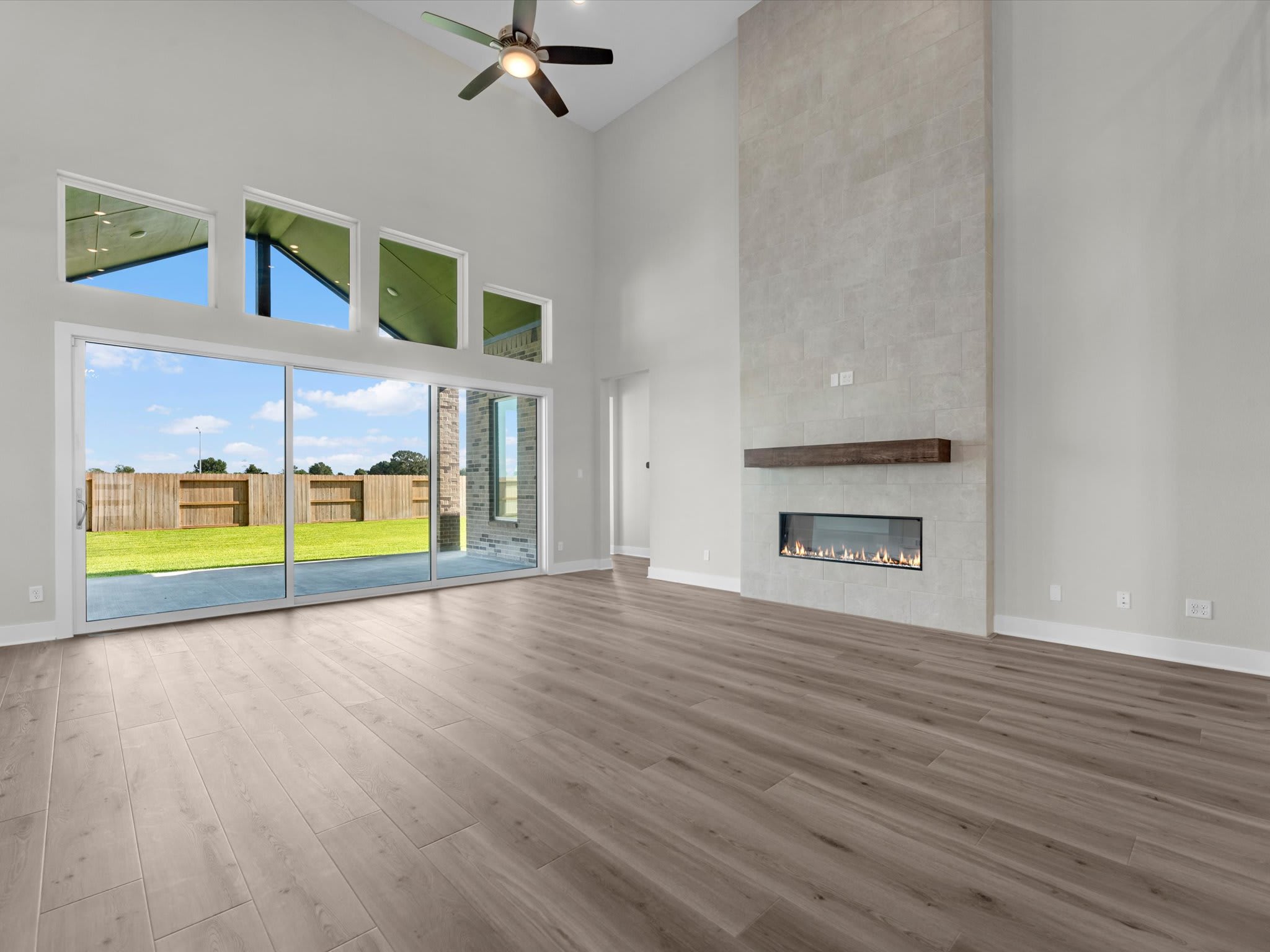 A spacious living room with light wood flooring and a modern fireplace in the foreground opens up to a sunny backyard through large sliding glass doors in the background.