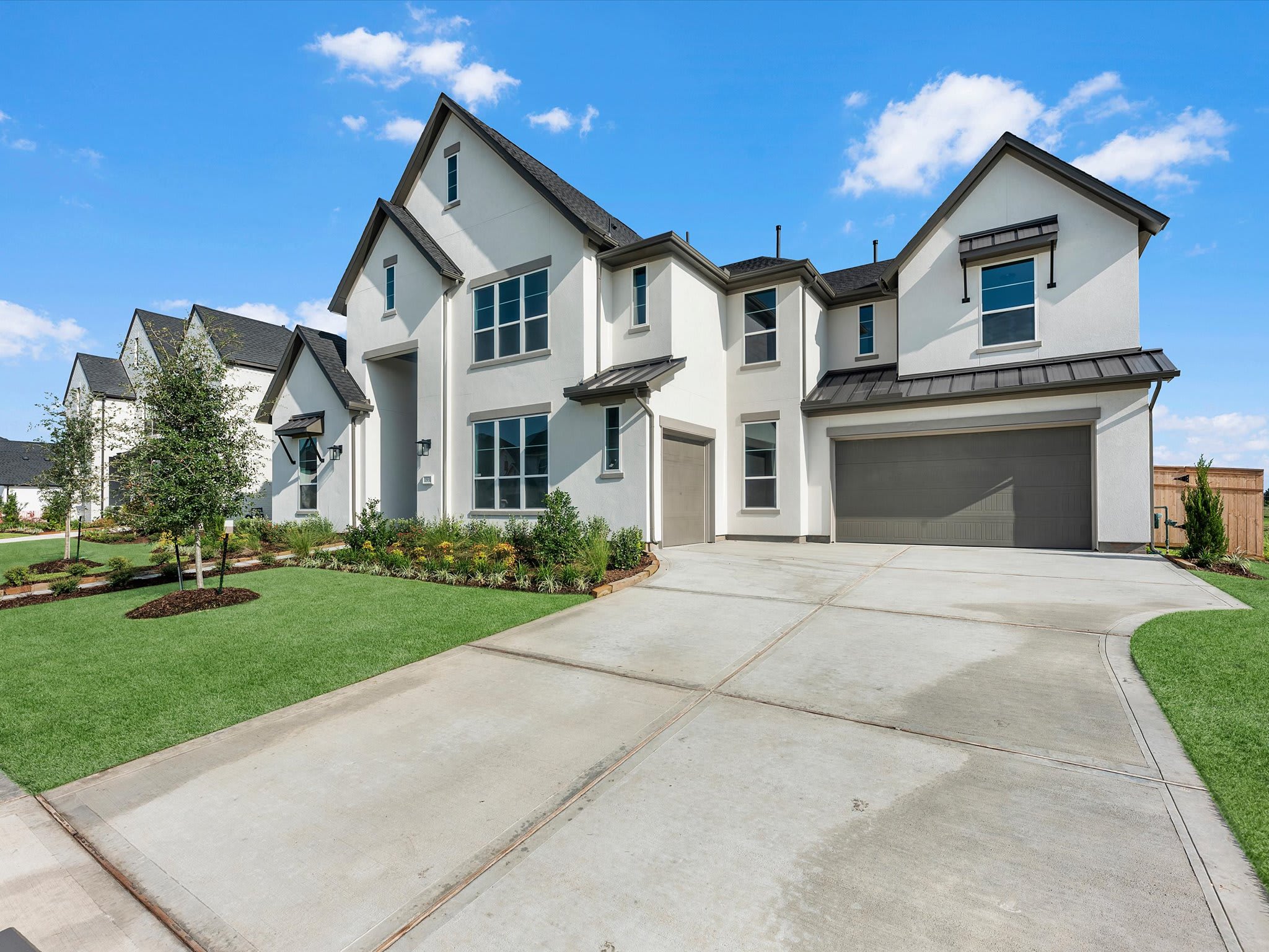 A large, modern white house with a grey garage and black roof stands behind a concrete driveway and a manicured green lawn under a bright blue sky.