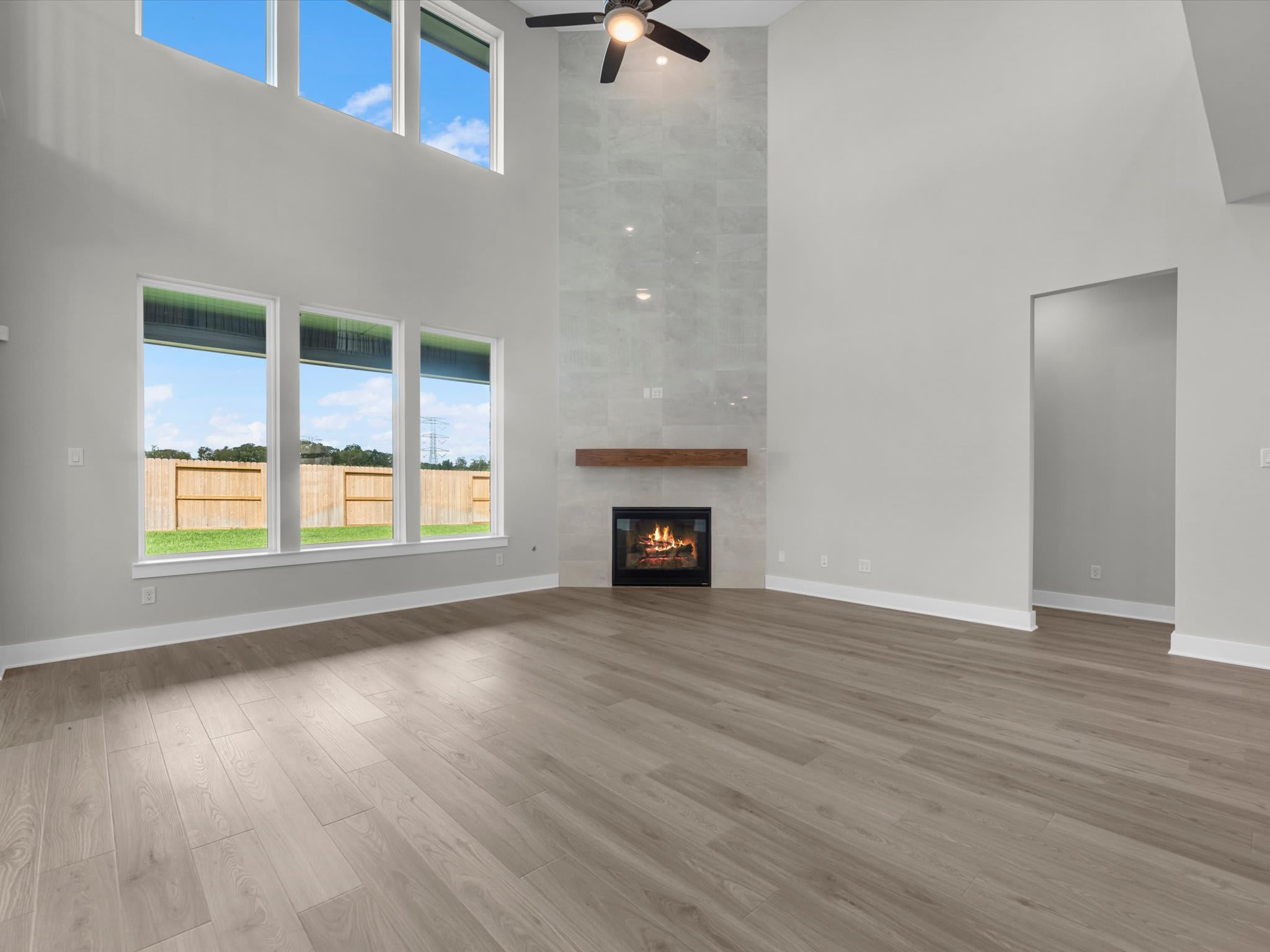 An empty living room with light wood floors features a fireplace with a wooden mantel against a white wall, with large windows in the background showing a green yard and blue sky.
