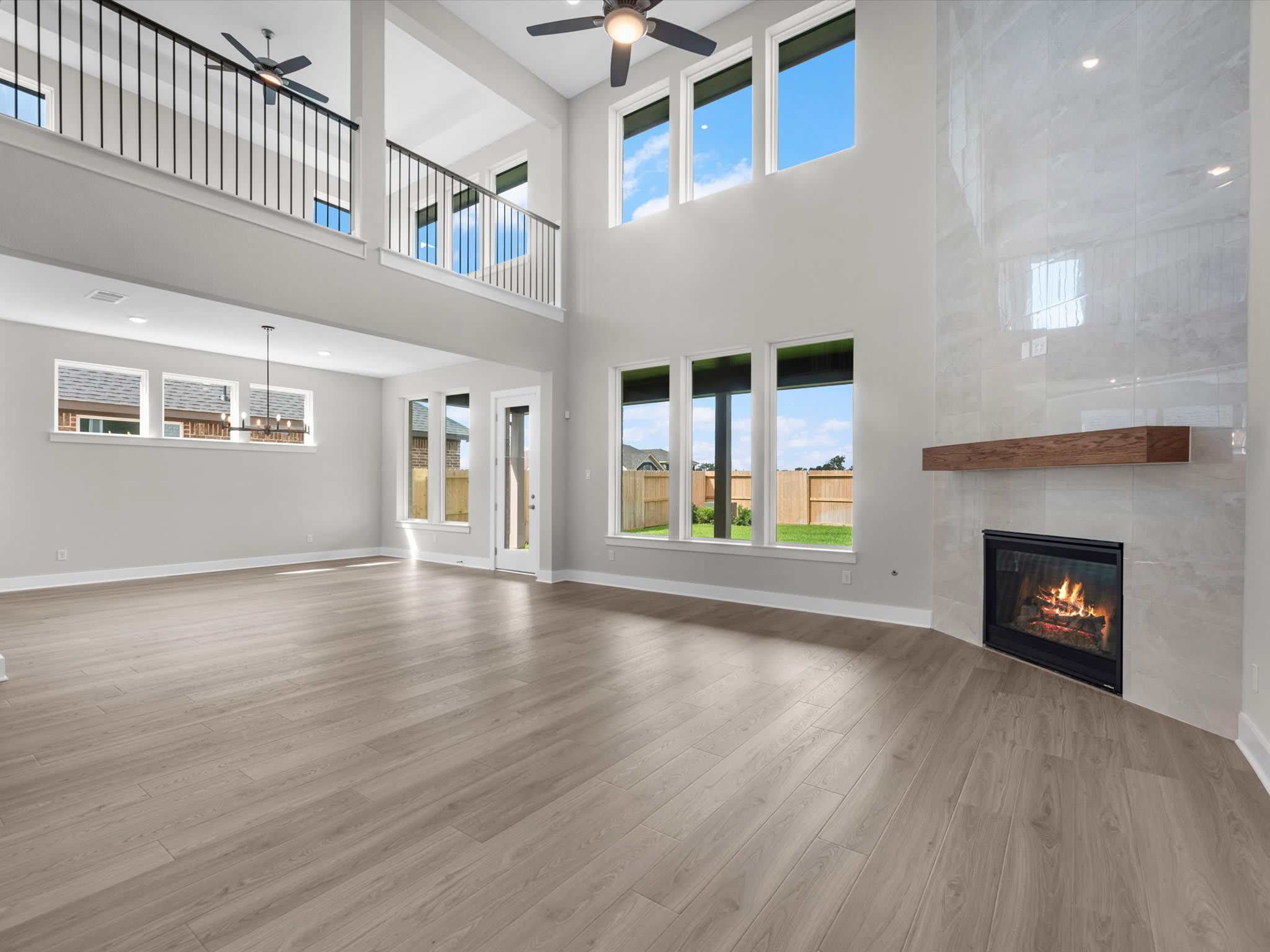 A spacious living room with light wood floors and a lit fireplace in the foreground opens up to a high-ceilinged area with a second-floor balcony and large windows in the background.